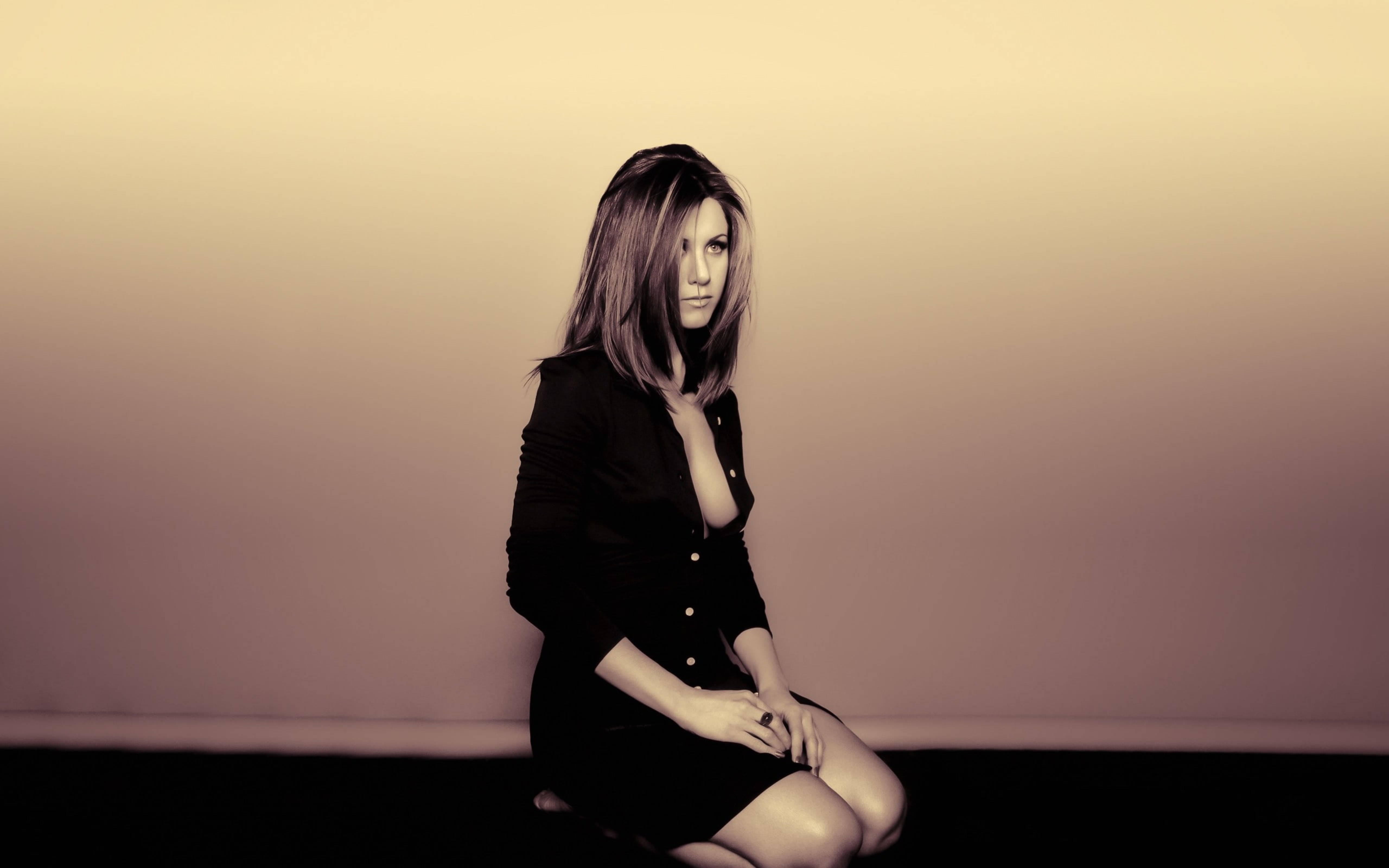 Jennifer Aniston 桌布 壁紙 — 一位身穿黑色連衣裙的女性跪在柔和照明的漸變牆前，表情沉思。