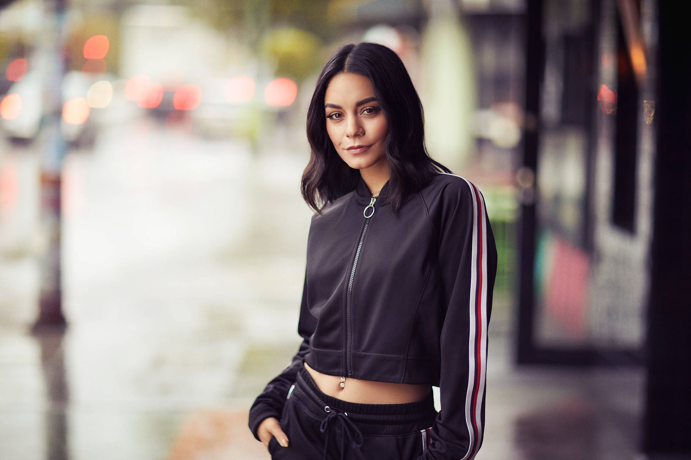 Vanessa Hudgens 桌布 壁紙 — 一位身穿黑色運動外套的年輕女性自信地站在濕漉漉的城市街道上，背景是模糊的霓虹燈。