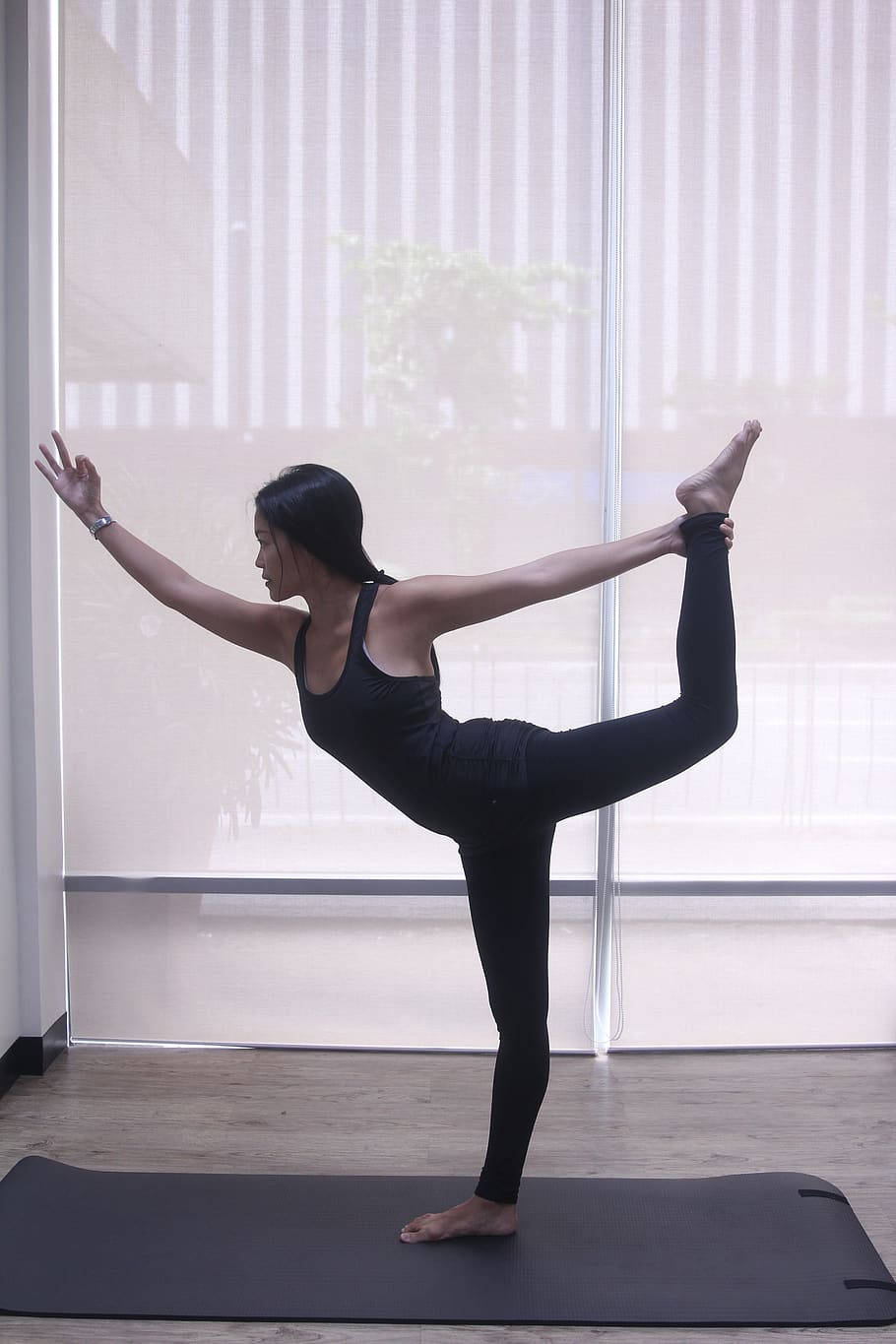 Exercise 桌布 壁紙 — 一位身穿黑色運動服的女士，在靠近大窗戶（配有薄紗百葉窗）的墊子上優雅地保持瑜伽姿勢平衡。