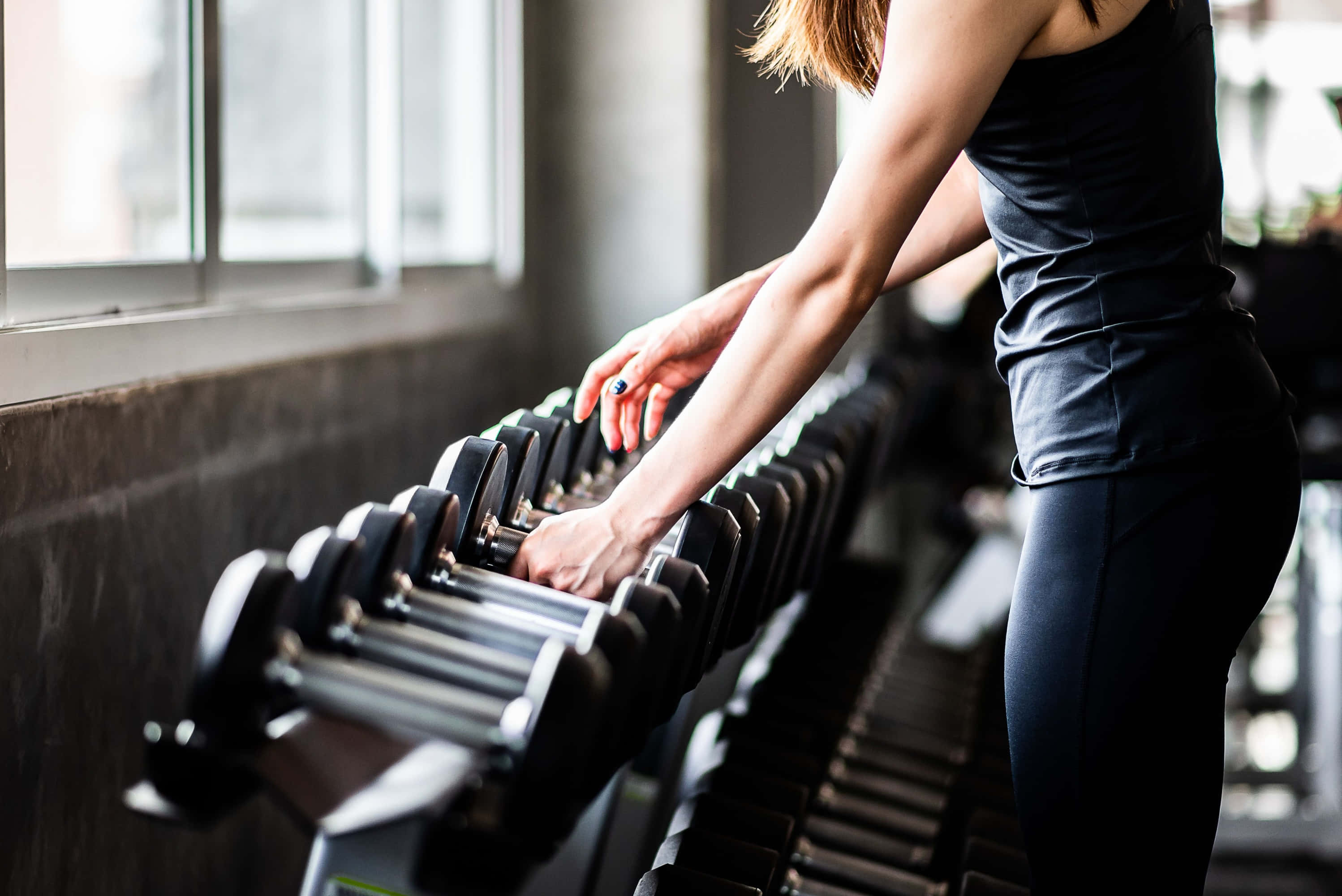 Fitness 桌布 壁紙 — 一位身穿黑色運動服的女士在一個有大型窗戶的現代健身房中從架子上挑選啞鈴。