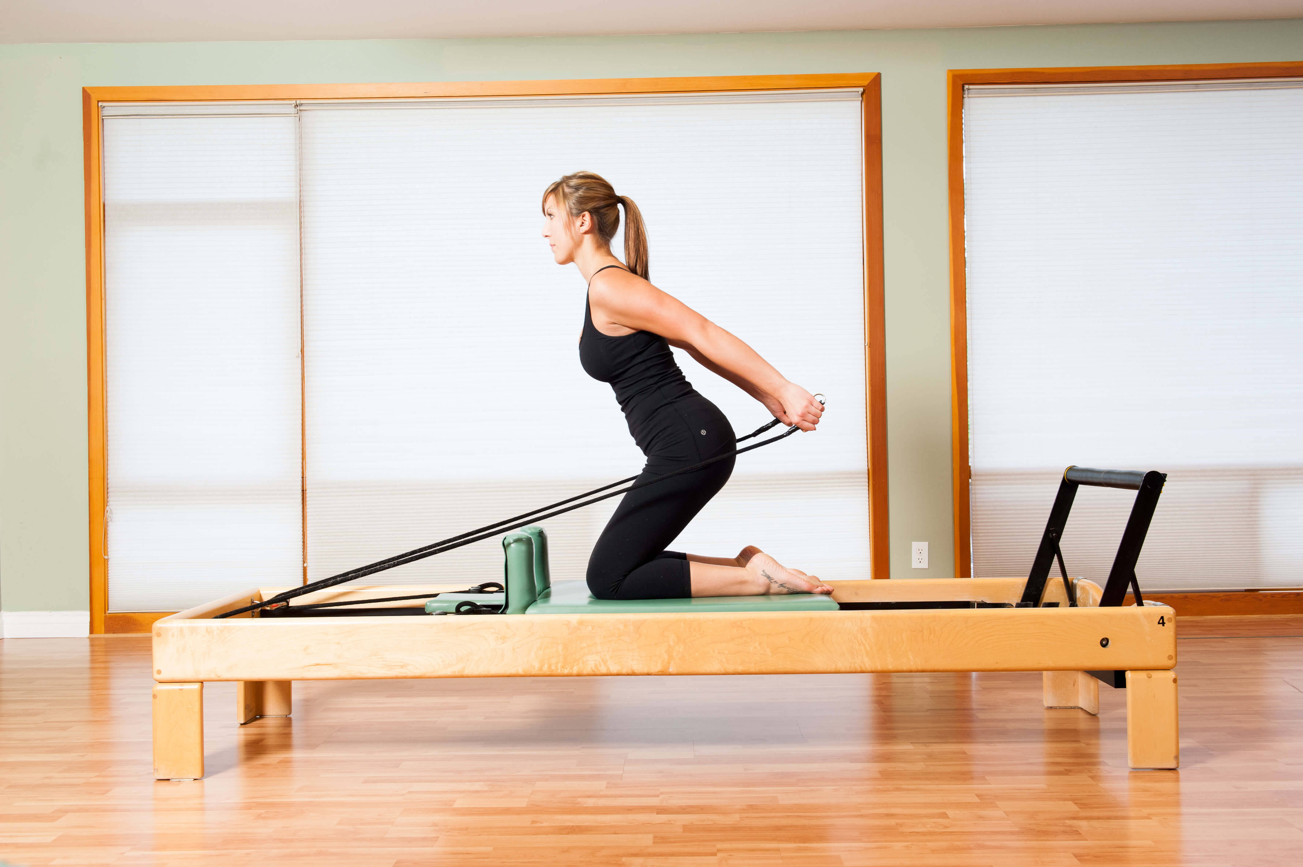 Exercise 桌布 壁紙 — 一位身穿黑色運動服的女士在明亮的舞蹈室裡，在木製的塑身機上進行皮拉提斯運動，室內有木地板和大窗戶。