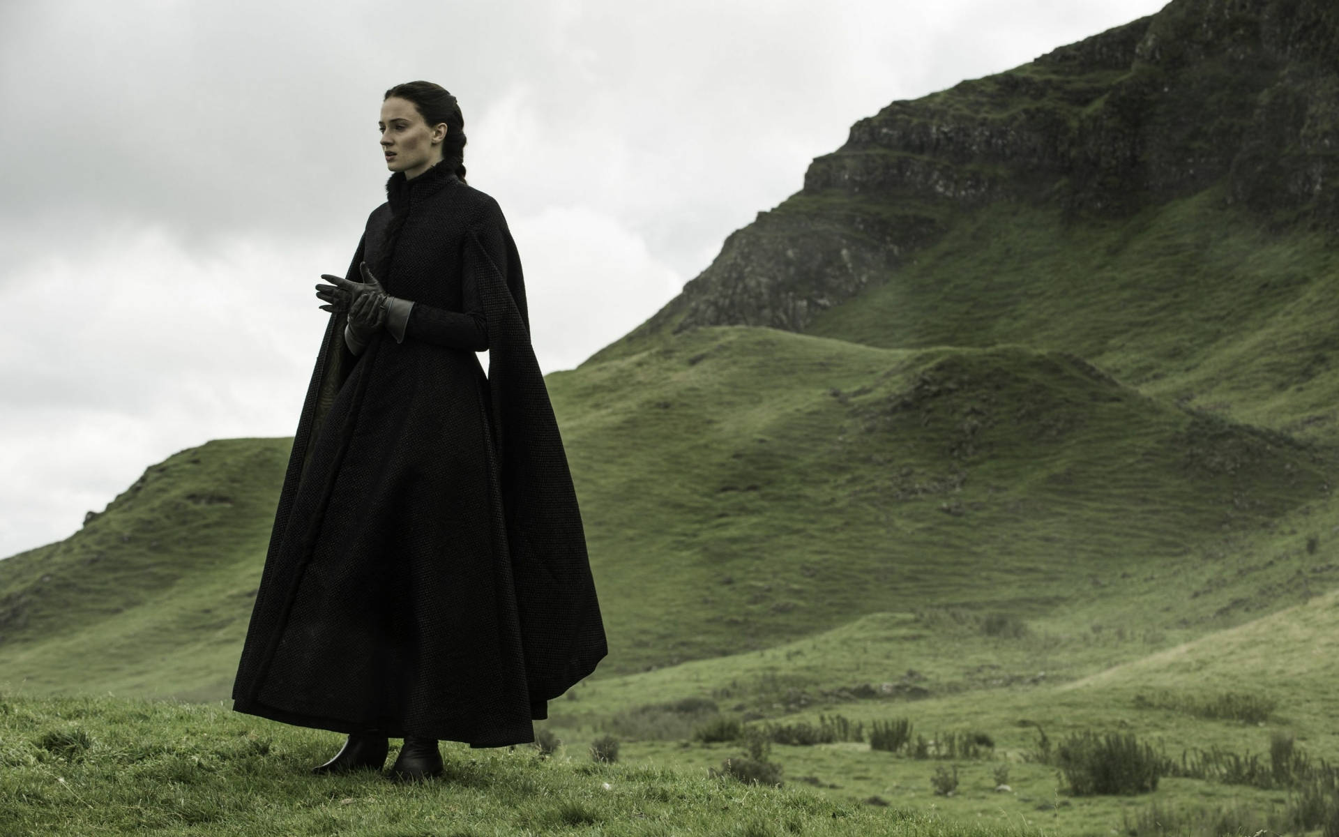 Sansa Stark 桌布 壁紙 — 一位身穿黑色長斗篷的女子獨自站在長滿青草的山坡上，天空陰沉，身後是連綿起伏的綠色山丘。