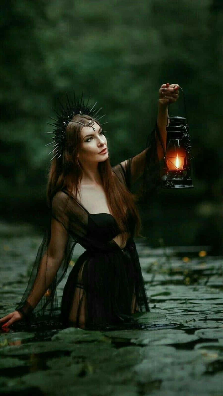 Witch Costumes 桌布 壁紙 — 一位身穿黑色長袍、頭戴尖刺王冠的女性站在黑暗森林的池塘中，手持發光燈籠。
