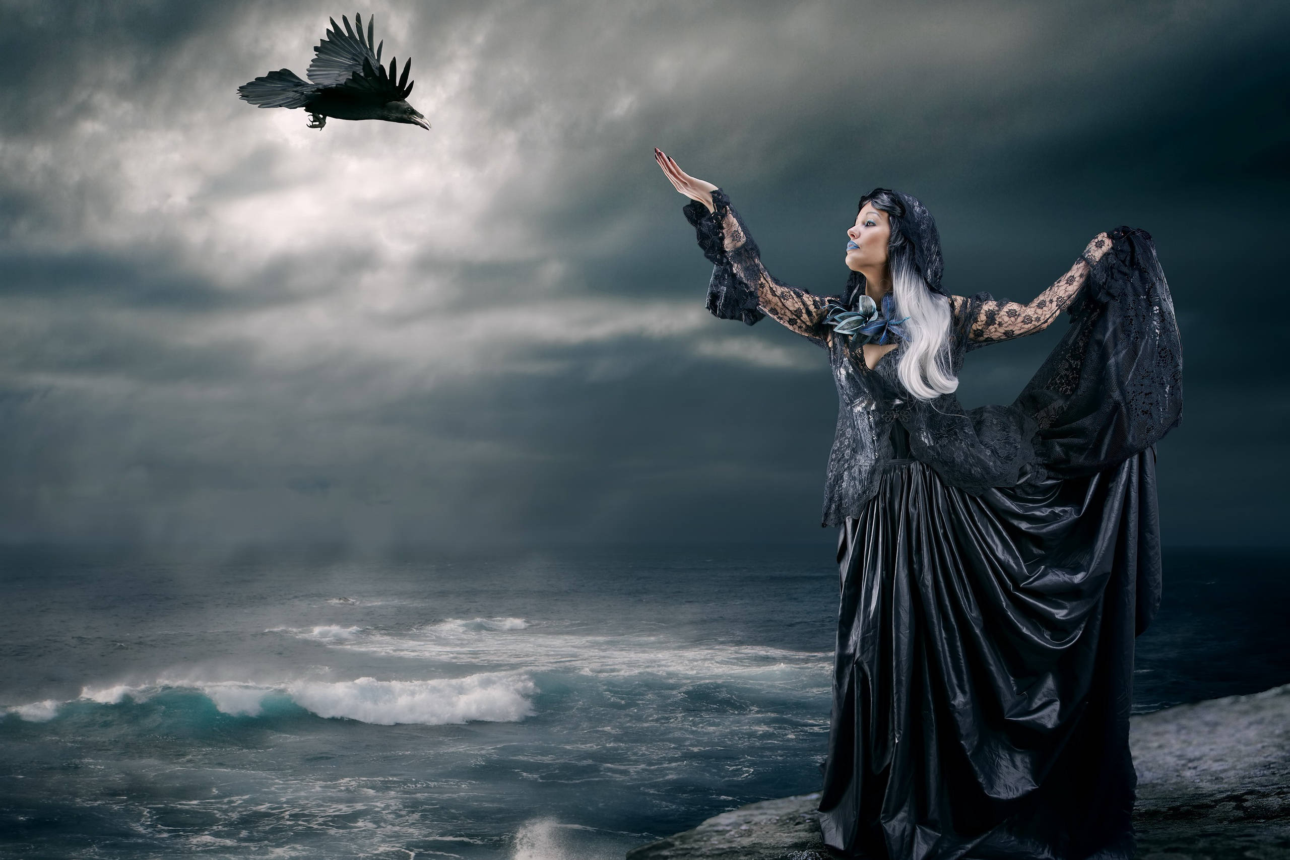 Goth 桌布 壁紙 — 一位身穿黑色長袍的女性站在風暴中的懸崖上，向著飛翔的烏鴉伸出手，背景是烏雲密布和翻騰的海浪。