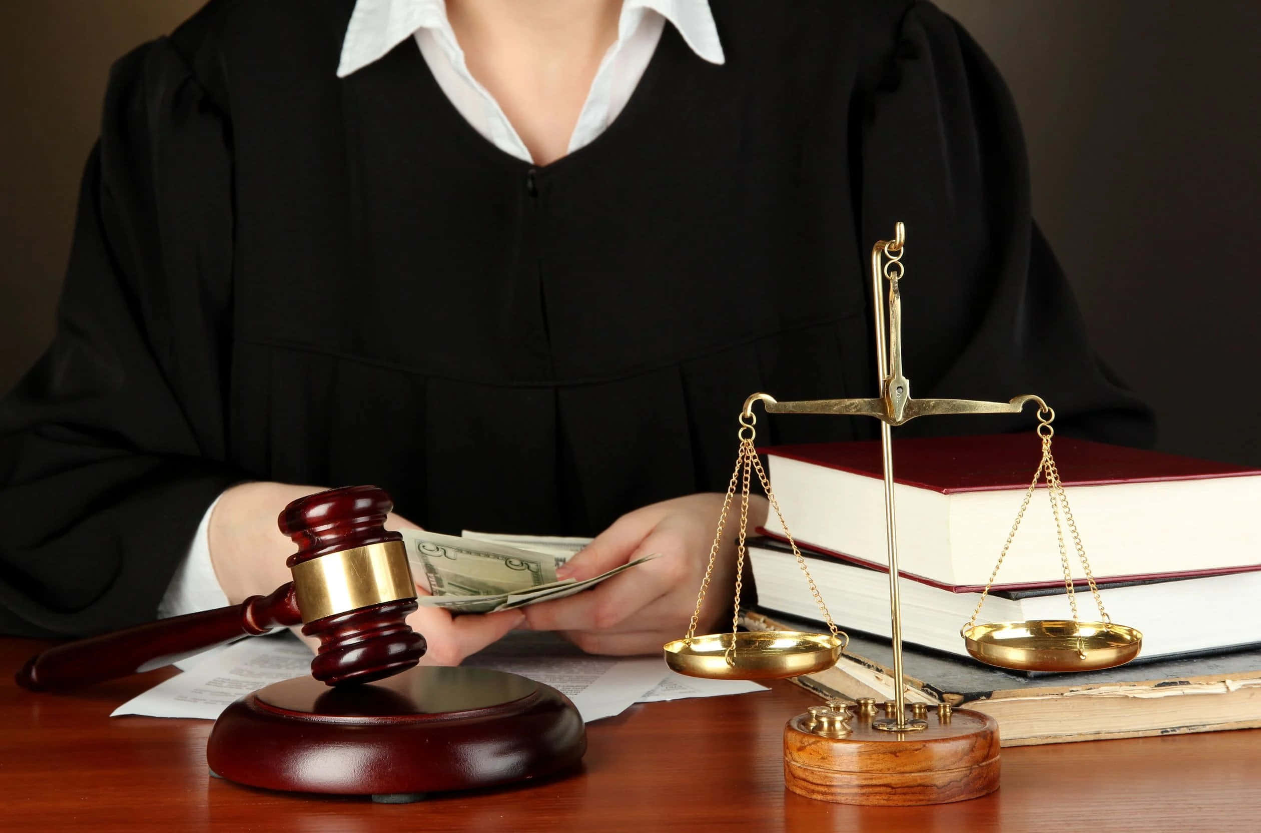 Law 桌布 壁紙 — 一位身穿黑色長袍的法官坐在木桌前，手持現金，旁邊放著法槌、正義天秤和法律書籍。