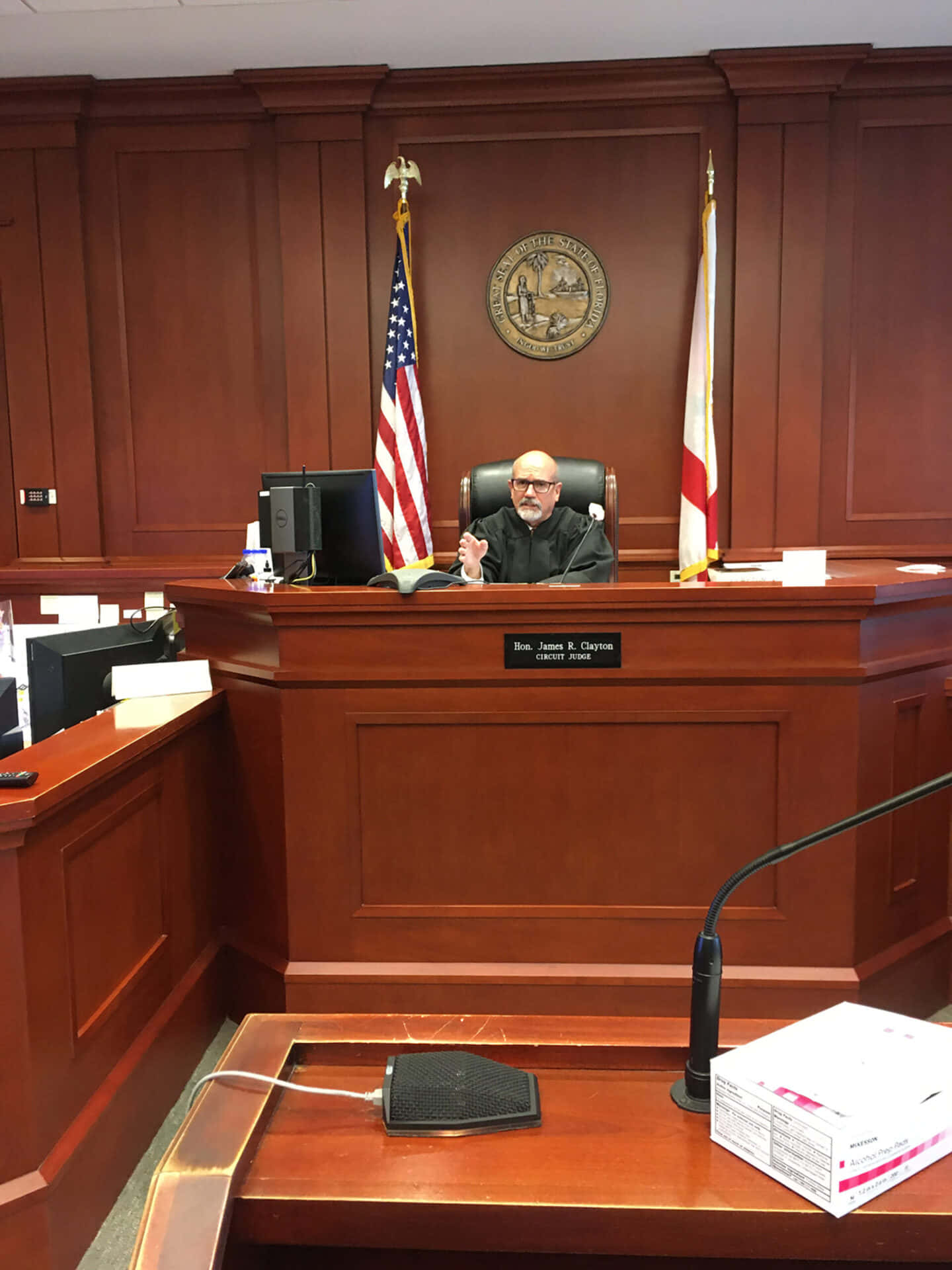 Courtroom 桌布 壁紙 — 一位身穿黑色長袍的法官坐在正式法庭的木製長椅上，牆上有美國和州旗幟以及印章。