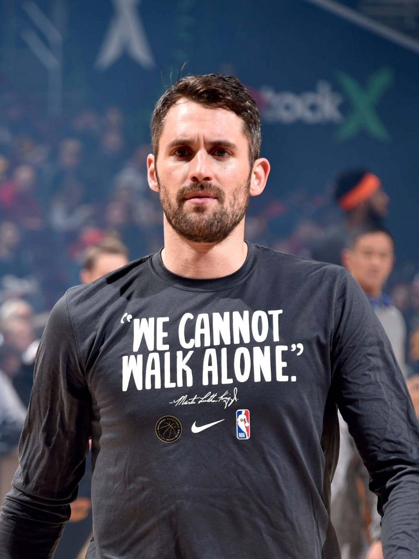 Kevin Love 桌布 壁紙 — 一位身穿黑色長袖衫、印有「我們不能獨自行走」語句的籃球運動員，站在擠滿觀眾的場館內。
