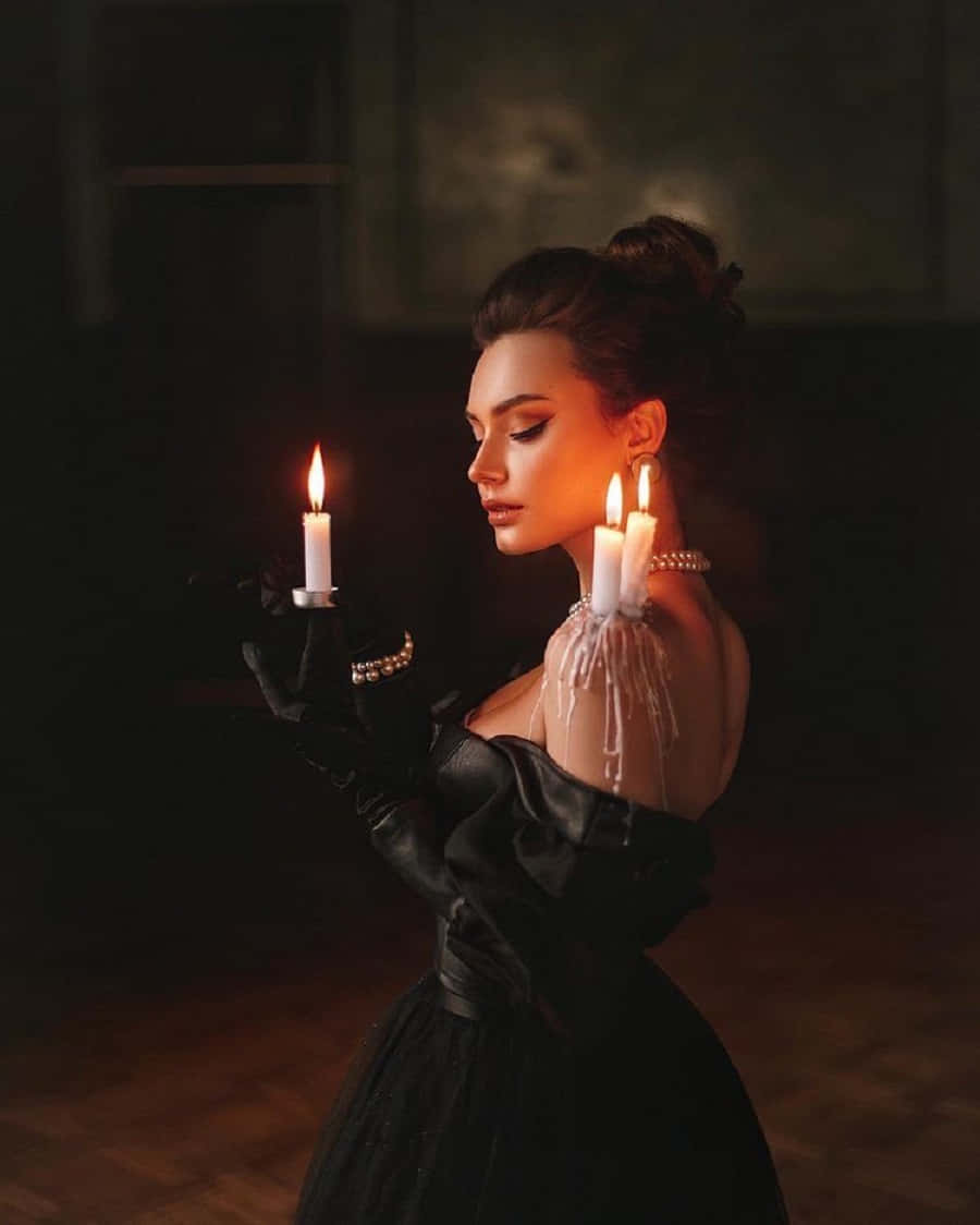 Witch Costumes 桌布 壁紙 — 一位身穿黑色長裙的女性在昏暗的房間裡手持點燃的蠟燭，溫暖的光芒照在她的臉上。
