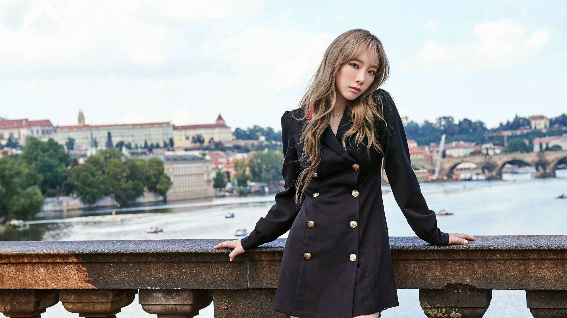 태연 桌布 壁紙 — 一位身穿黑色雙排扣大衣的女子倚在石欄杆上，俯瞰著一條河流，河上有歷史悠久的橋樑和建築，天空部分多雲。