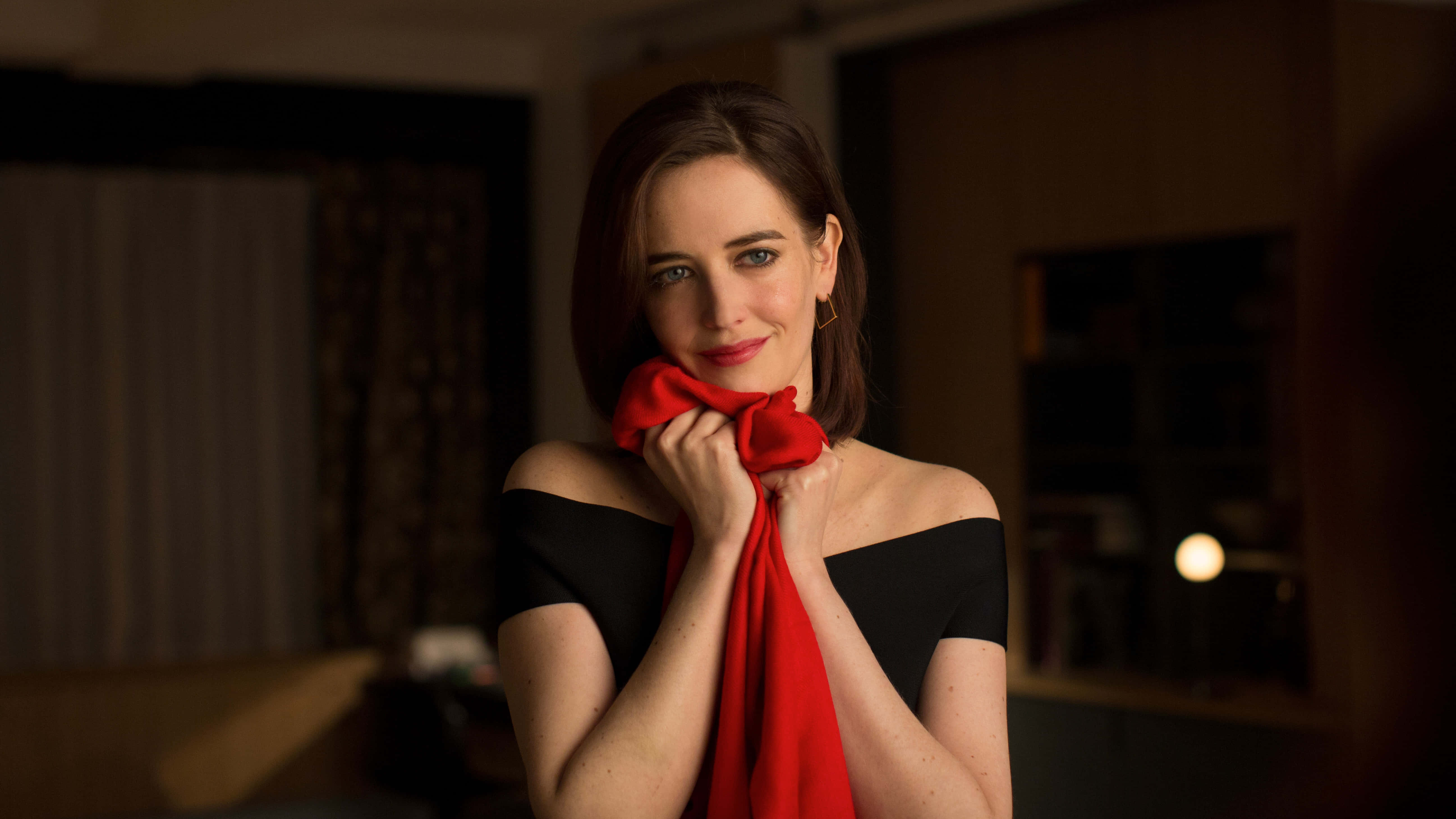 eva green 桌布 壁紙 — 一位身穿黑色露肩連衣裙的女性在昏暗的房間裡將一條鮮紅色的圍巾貼在脖子上。