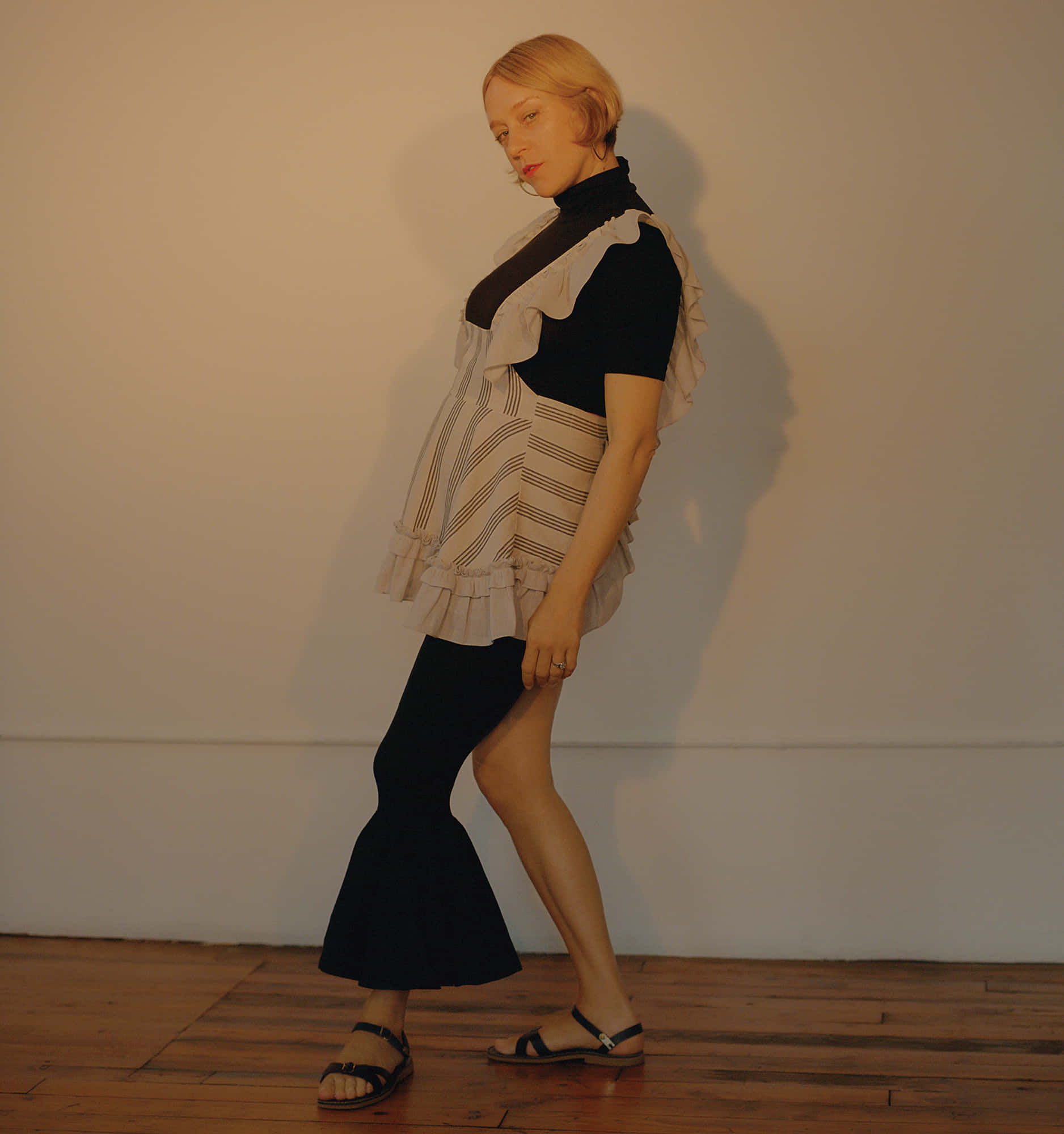 Chloë Sevigny 桌布 壁紙 — 一位身穿黑色高領毛衣和條紋荷葉邊背心的女性，站在一個光線溫暖、有木地板和素色牆壁的房間裡。