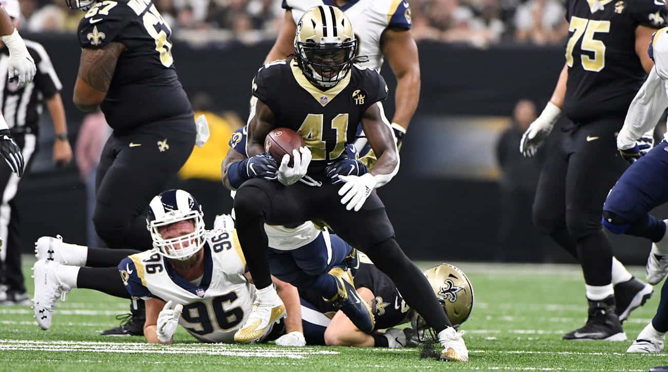 Alvin Kamara 桌布 壁紙 — 一位身穿黑金色球衣的紐奧良聖徒球員帶著美式足球奔跑，同時被洛杉磯公羊的防守球員在場上擒抱。
