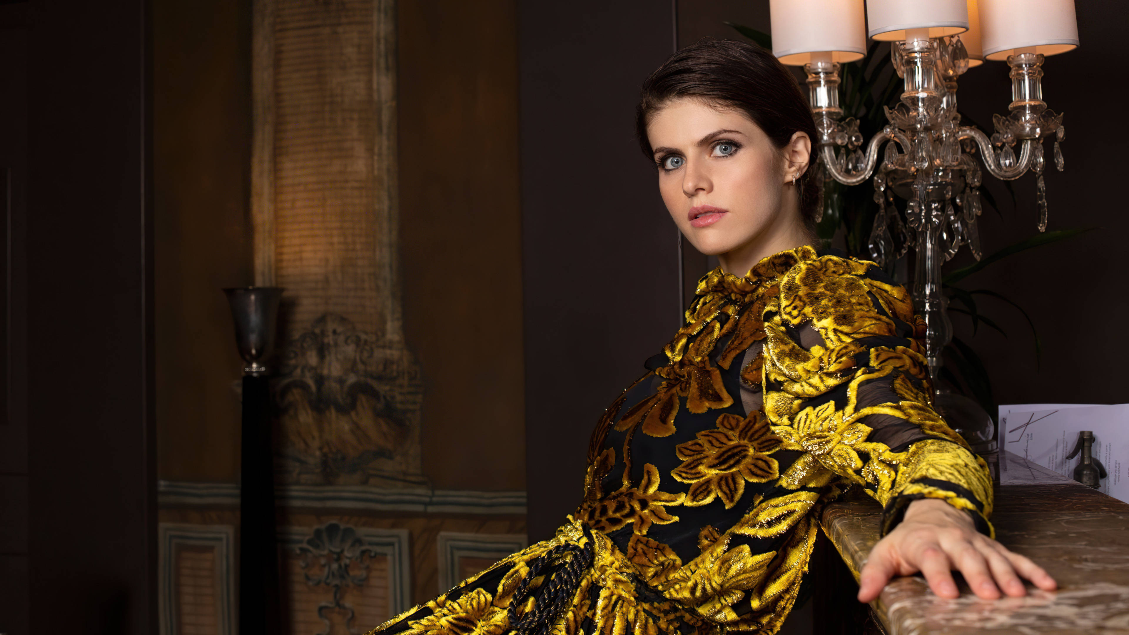 Alexandra Daddario 4K 桌布 壁紙 — 一位身穿黑金花卉長裙的女性，在裝飾華麗的房間裡，優雅地在水晶吊燈旁擺姿勢。