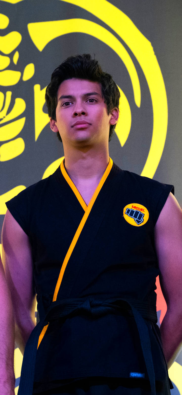 Man Standing 桌布 壁紙 — 一位身穿黑黃色武術服的年輕男子自信地站在醒目的黃色圓形標誌背景前。