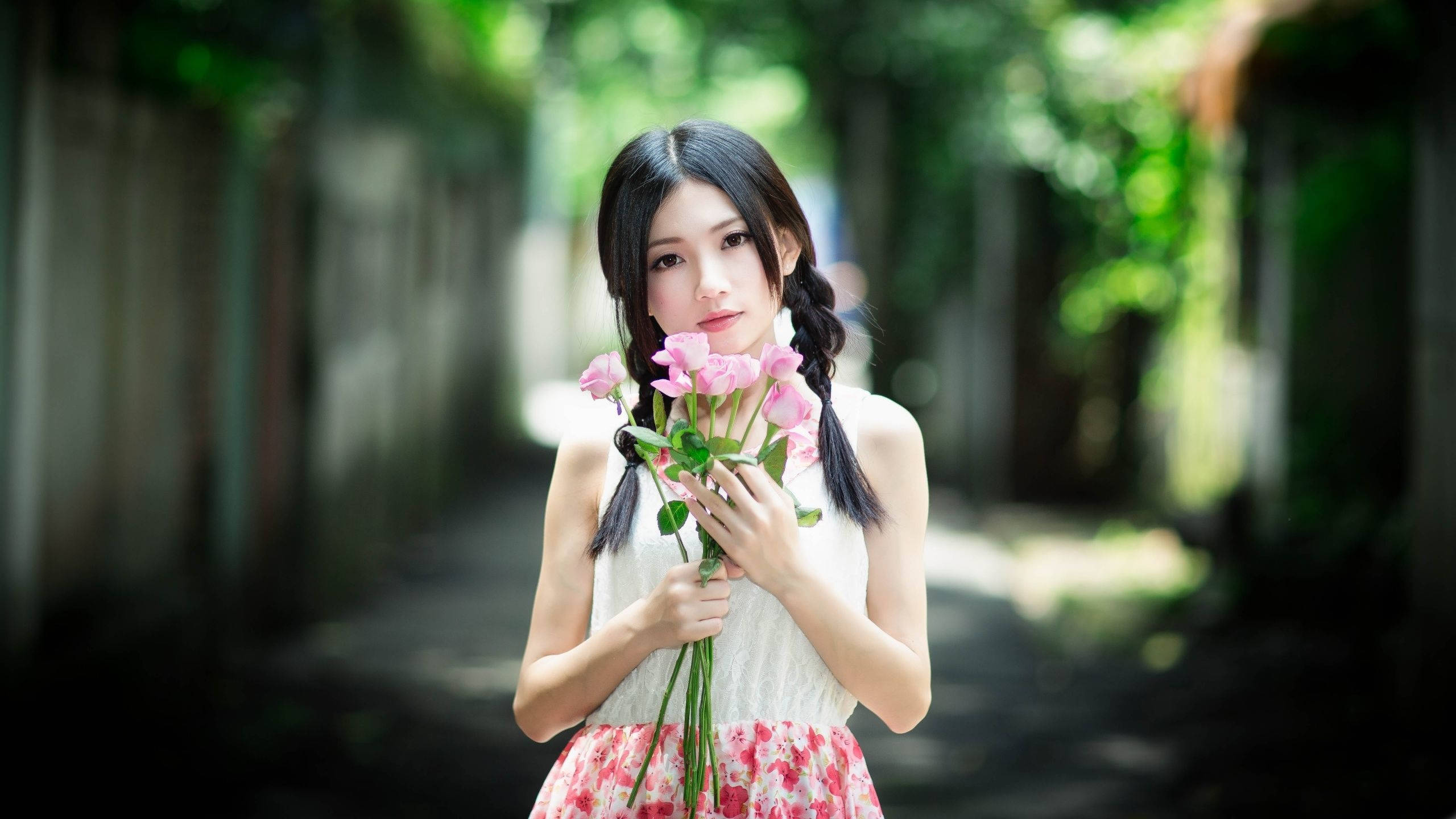 美女 桌布 壁紙 — 一位辮子年輕女子在陽光明媚的花園小徑上手持粉紅玫瑰，四周環繞著茂密的綠色樹葉。