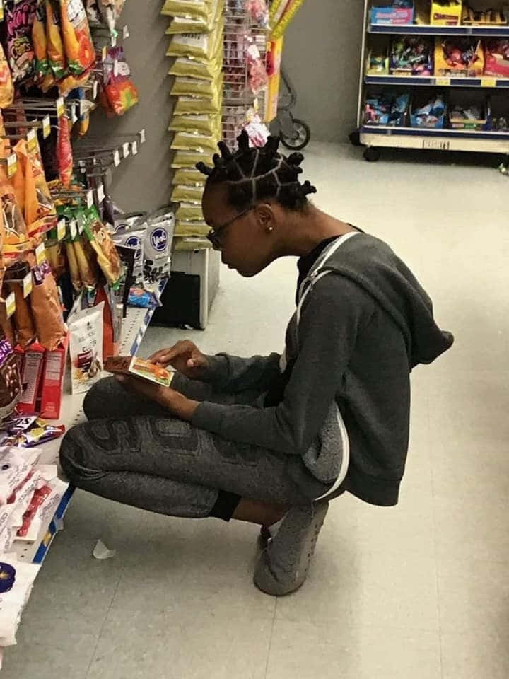 搞笑 桌布 壁紙 — 一位辮子年輕女子蹲在雜貨店貨架通道，檢查一個被色彩繽紛包裝商品包圍的零食盒。