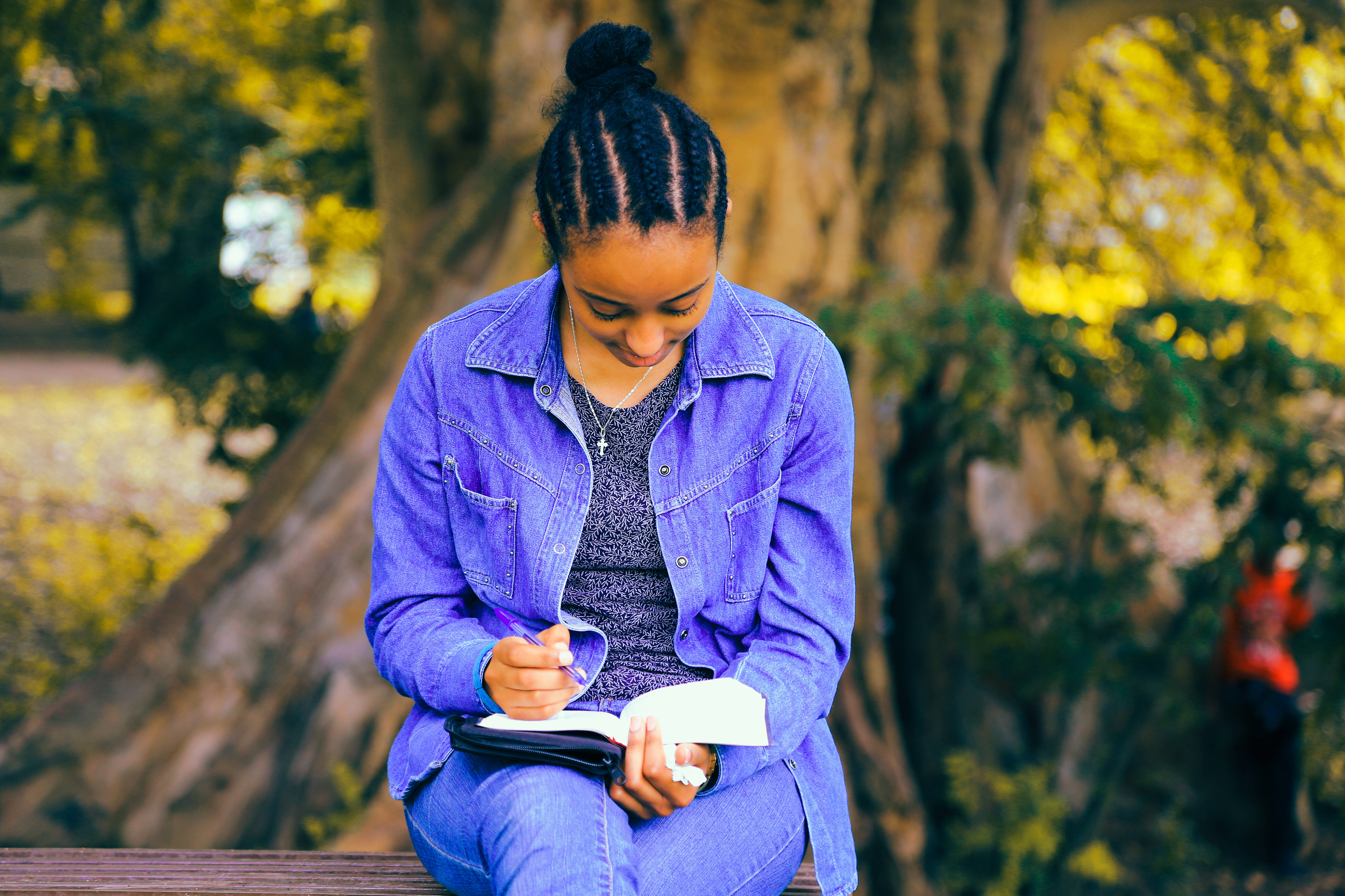 看書 桌布 壁紙 — 一位辮子年輕女性坐在陽光普照的公園長椅上，在金黃色的秋樹下寫筆記。