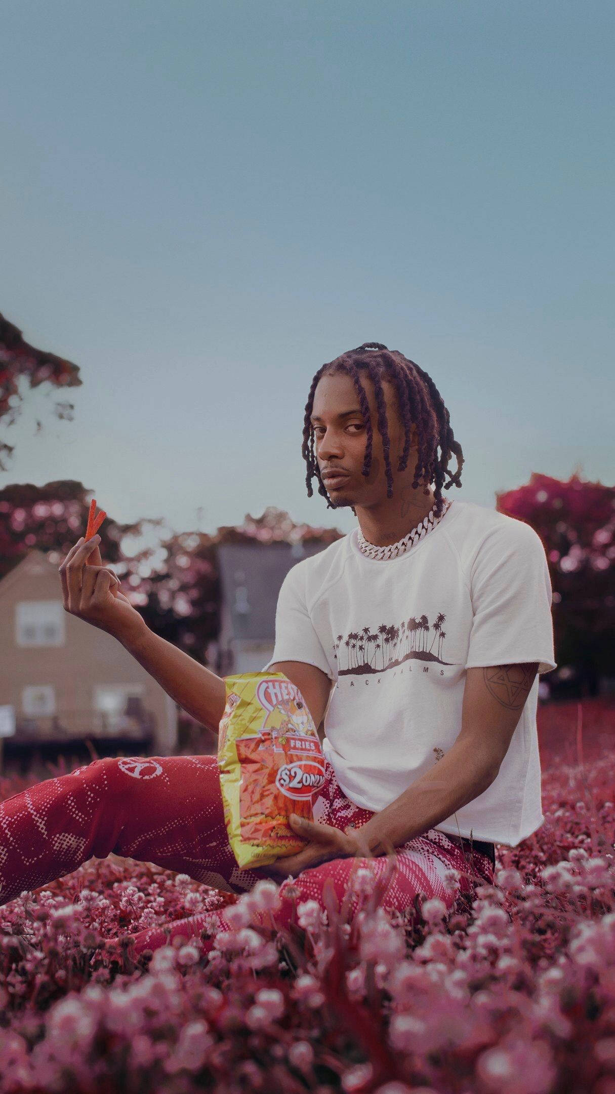 Rappers 桌布 壁紙 — 一位辮子年輕男子坐在粉紅色花田裡，拿著一包洋芋片和一片洋芋片，穿著紅色褲子和印有棕櫚樹圖案的白色T恤。