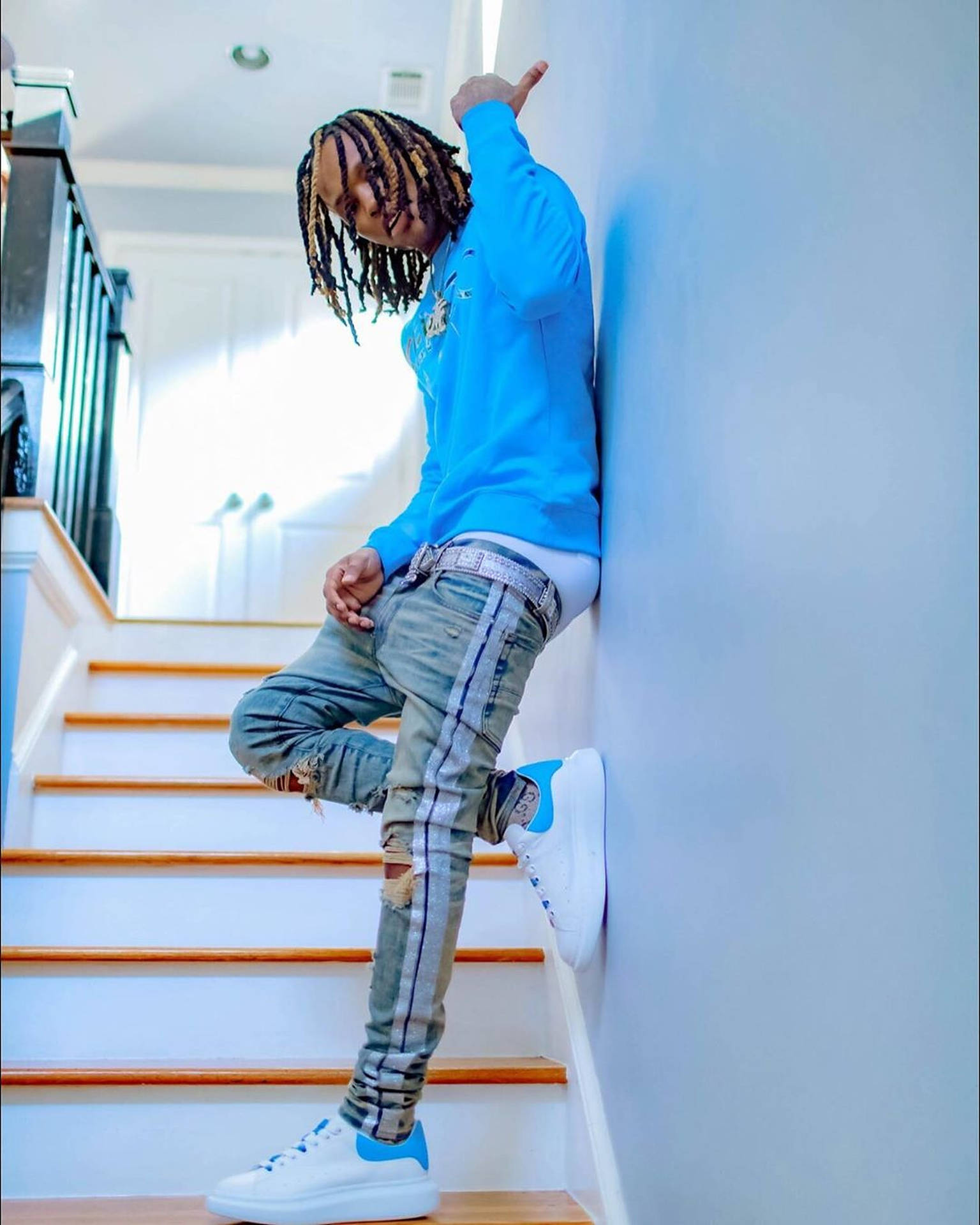 穿著藍色運動衫和破洞牛仔褲的年輕男子靠在樓梯旁的牆上。 — King Von 壁紙