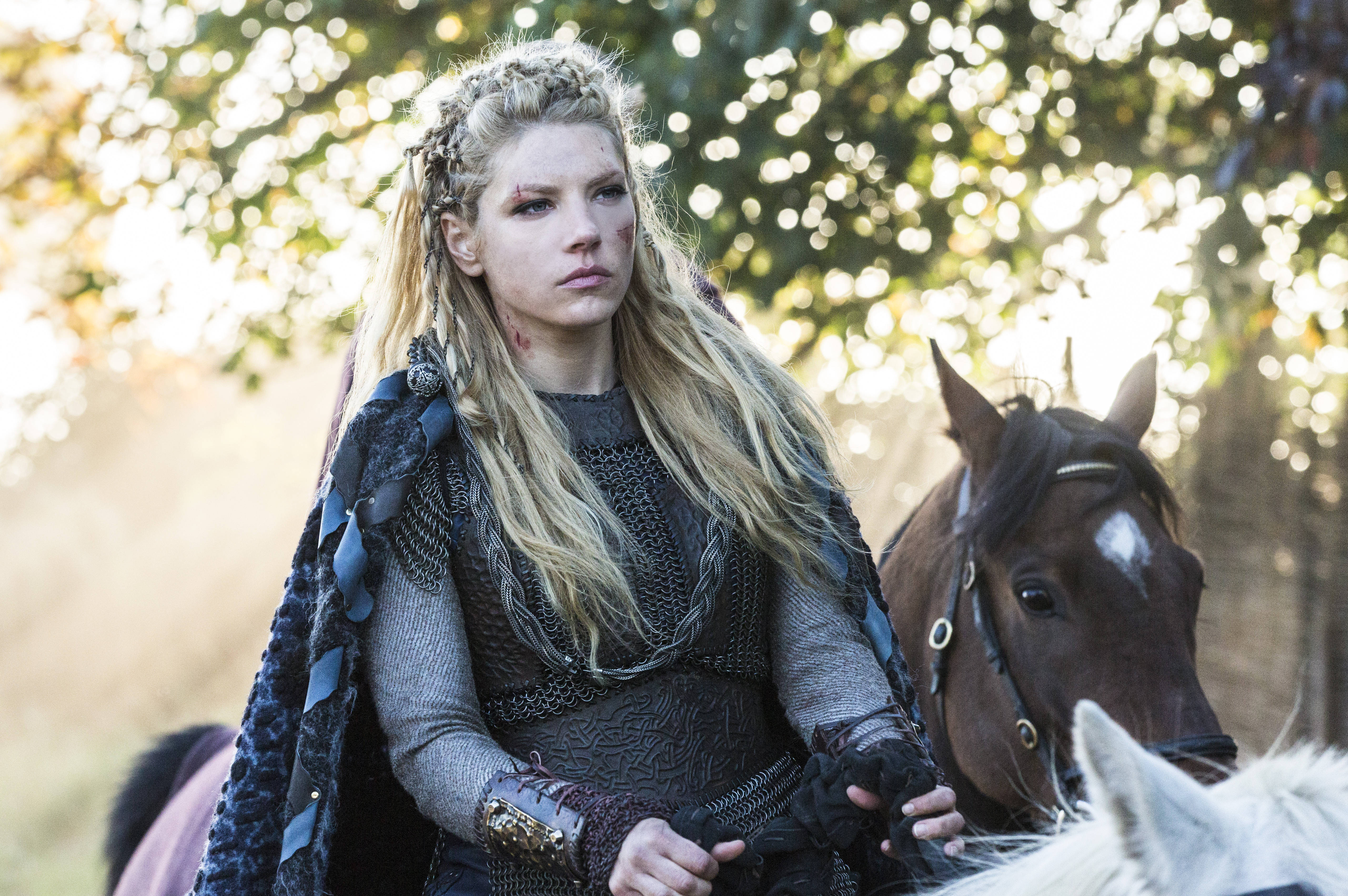 Vikings 桌布 壁紙 — 一位辮子金髮、帶有戰鬥傷疤的女戰士，騎著一匹黑馬穿過陽光明媚的秋日森林。