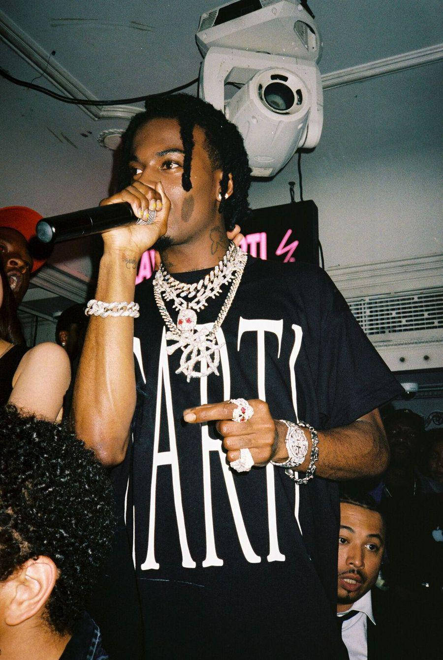 Carti 桌布 壁紙 — 一位辮子頭、戴鑽石鏈的饒舌歌手在昏暗俱樂部的舞台燈光下對著麥克風表演。
