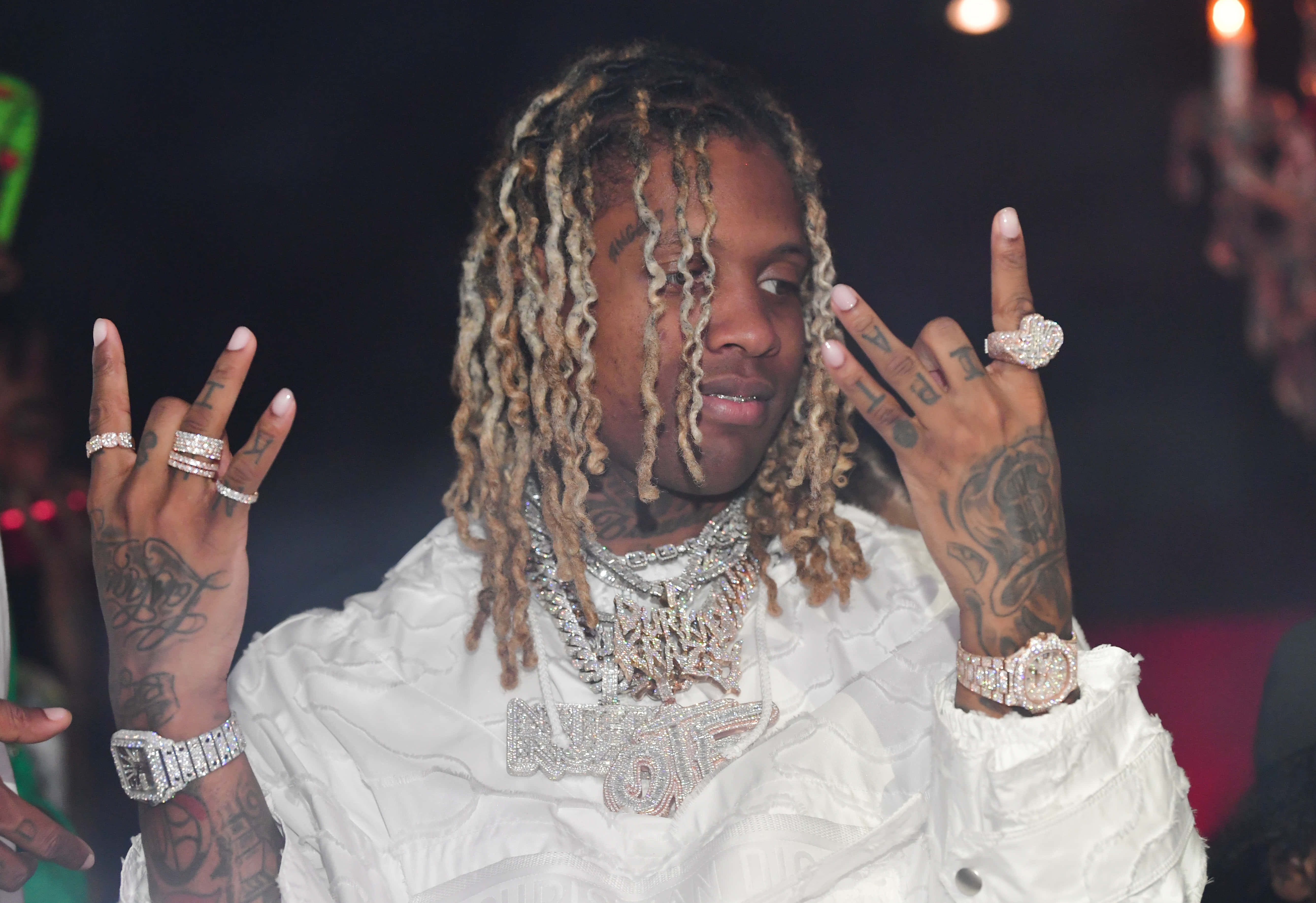 Lil Durk 桌布 壁紙 — 一位辮子頭和刺青的說唱歌手，在昏暗的俱樂部燈光下穿著白色服裝，閃耀著鑲鑽戒指和鏈條。