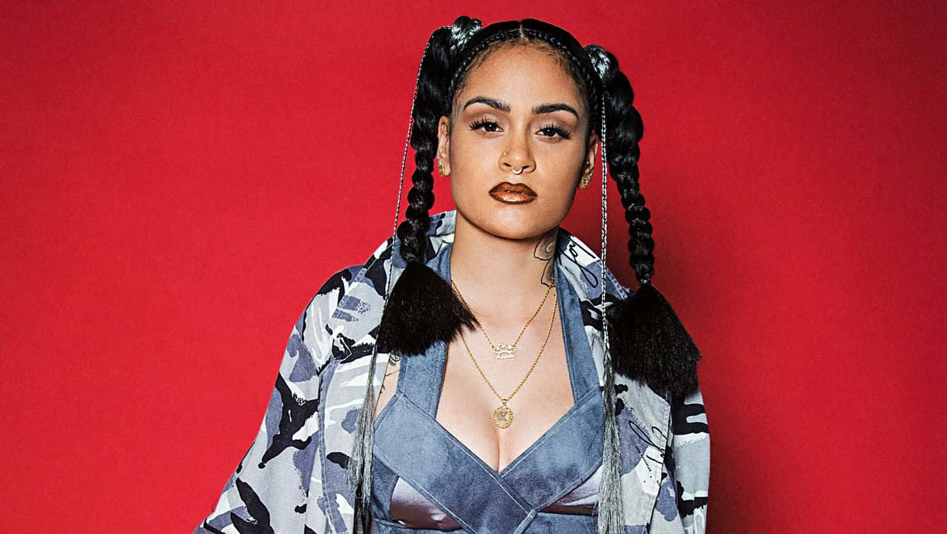 kehlani 桌布 壁紙 — 一位辮子頭髮、妝容大膽的女性，在鮮紅色背景前自信地擺姿勢，穿著圖案外套。