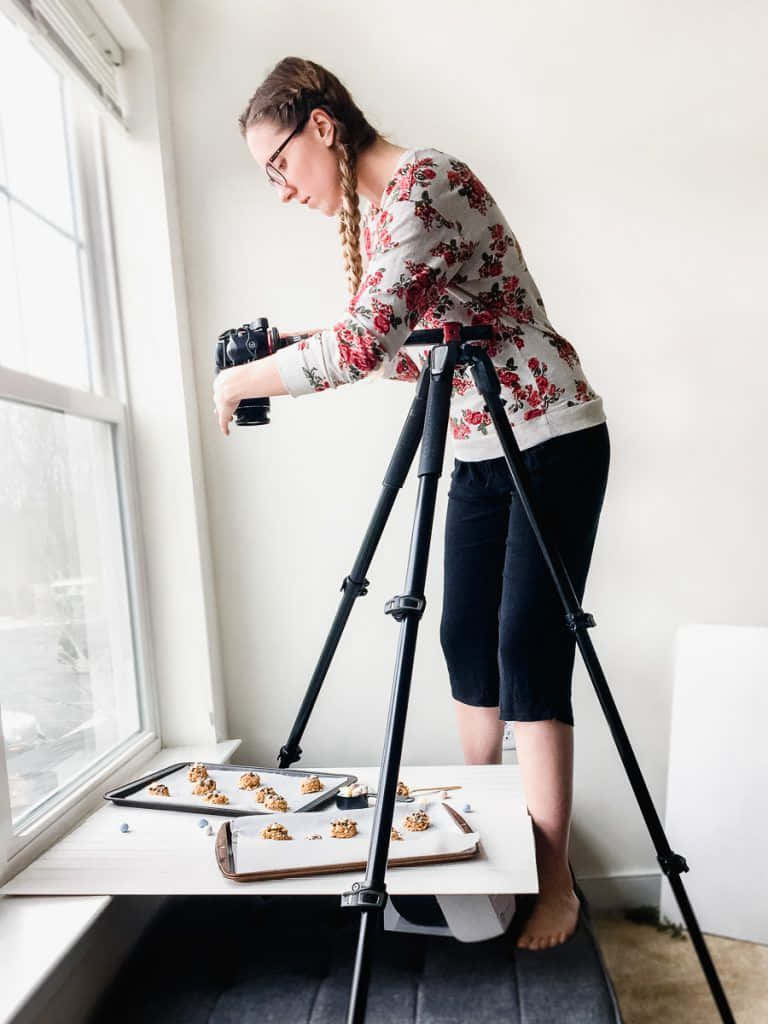 Holding Camera 桌布 壁紙 — 一位辮子頭髮、戴眼鏡的年輕女子俯身在三腳架上，拍攝明亮窗邊烤盤上擺放的餅乾。