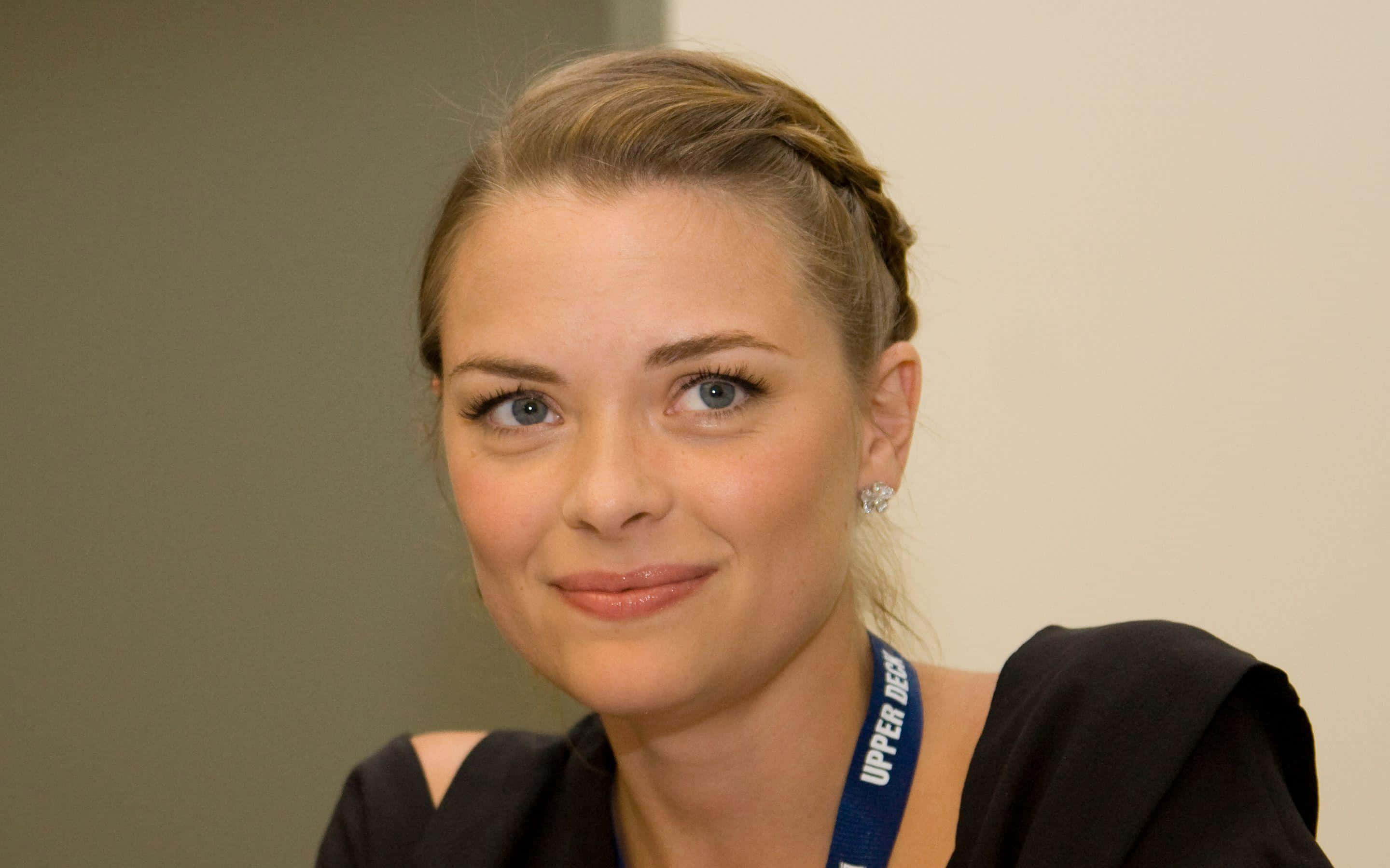 Jaime King 桌布 壁紙 — 一位辮子頭髮、掛著掛繩的女性在室內中性環境中對著鏡頭溫柔微笑。