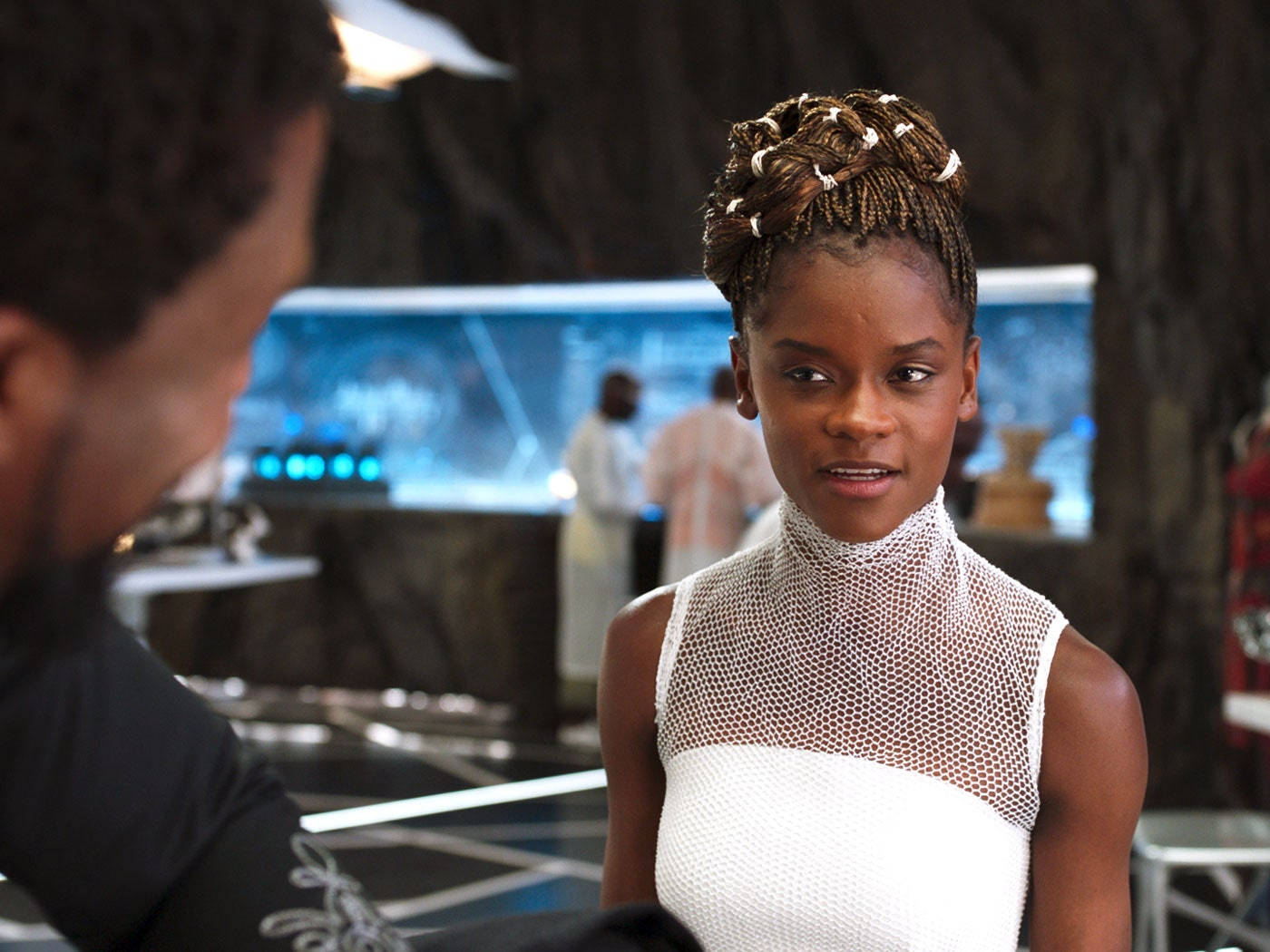 Shuri 桌布 壁紙 — 一位辮子頭髮、身穿白色網眼上衣的女性，在一個高科技的未來環境中專注地凝視著一名男子。