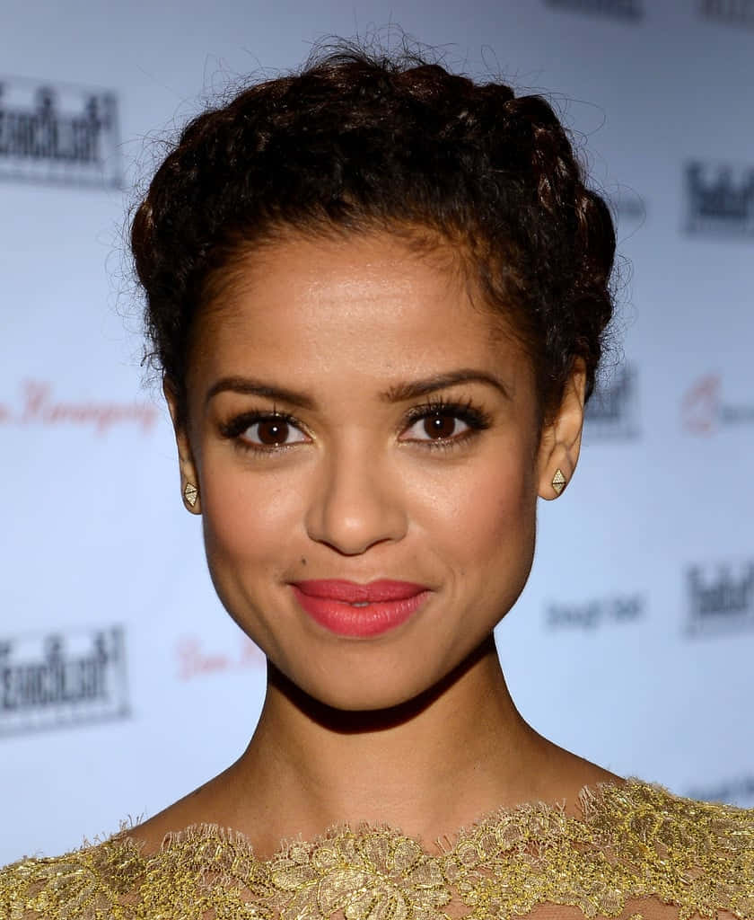 Gugu Mbatha-raw 桌布 壁紙 — 一位辮子頭髮、身穿金色蕾絲服飾的女性，在品牌背景前微笑面對鏡頭的近距離肖像。