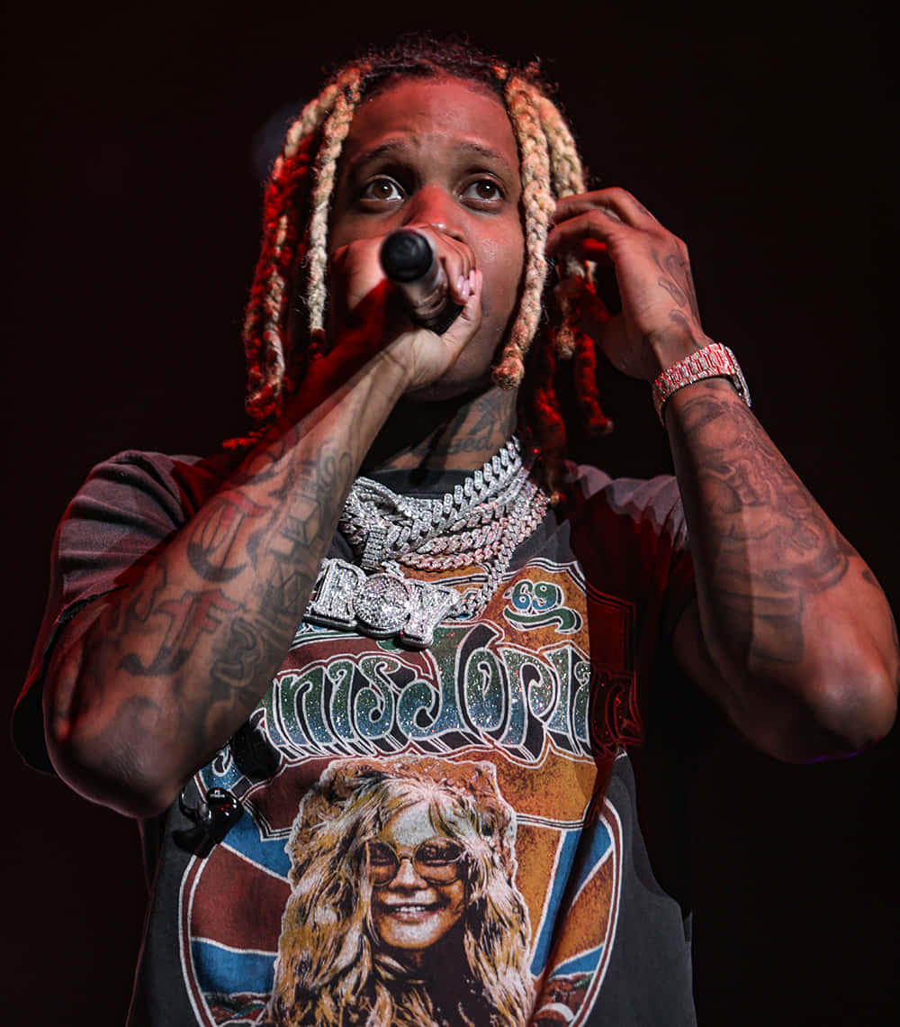 Lil Durk 桌布 壁紙 — 一位辮子頭髮和刺青的說唱歌手在紅色舞台燈光下拿著麥克風在舞台上表演。