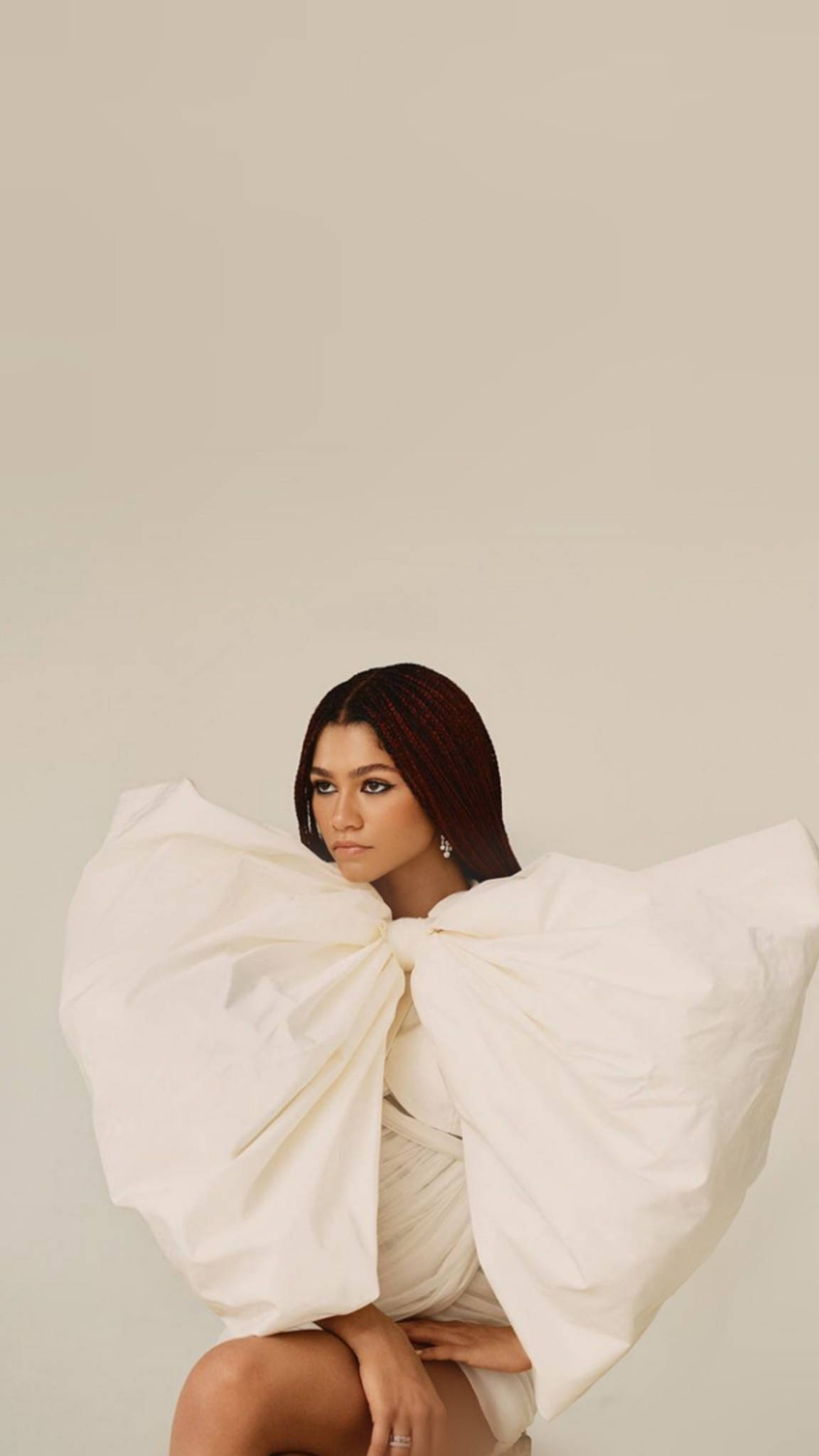 Zendaya 桌布 壁紙 — 一位辮子頭髮的女性，穿著白色大蝴蝶結連衣裙，平靜地坐在純米色背景前。