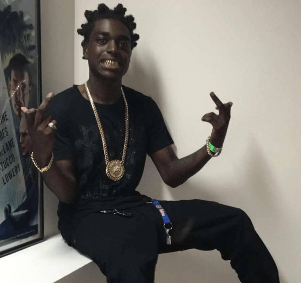 Kodak 桌布 壁紙 — 一位辮子頭髮的年輕男子自信地坐在白色邊緣上，戴著金鏈，露出笑容並做手勢。