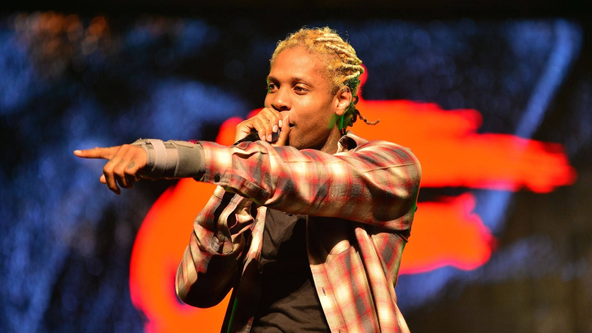 Lil Durk 桌布 壁紙 — 一位辮子頭髮的男性表演者穿著格紋襯衫，在充滿活力的舞台燈光下指向前方，同時對著麥克風唱歌。