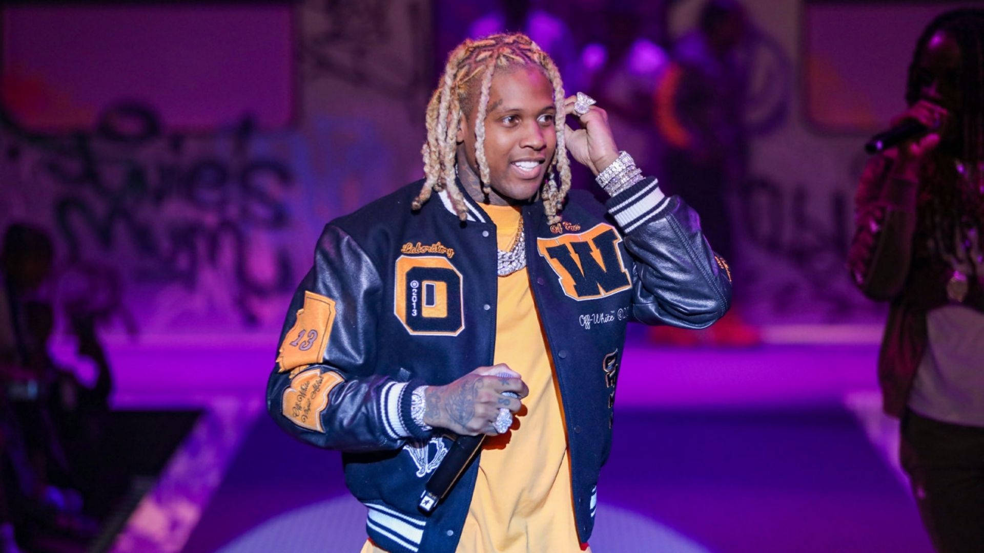 Lil Durk 桌布 壁紙 — 一位辮子頭髮的饒舌歌手在紫色燈光、塗鴉背景的舞台上微笑著調整耳朵。