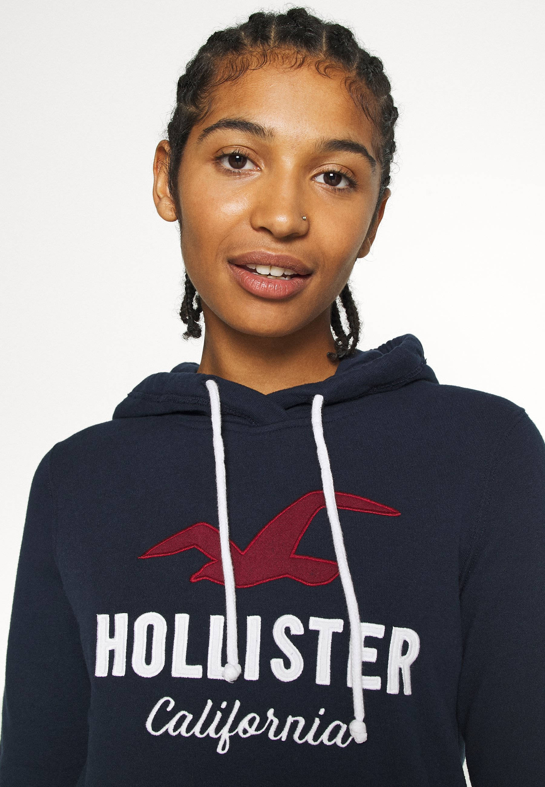 Hollister 桌布 壁紙 — 一位辮子髮型的年輕女性，在純白色背景前穿著帶有紅色標誌和白色抽繩的深藍色Hollister連帽衫。