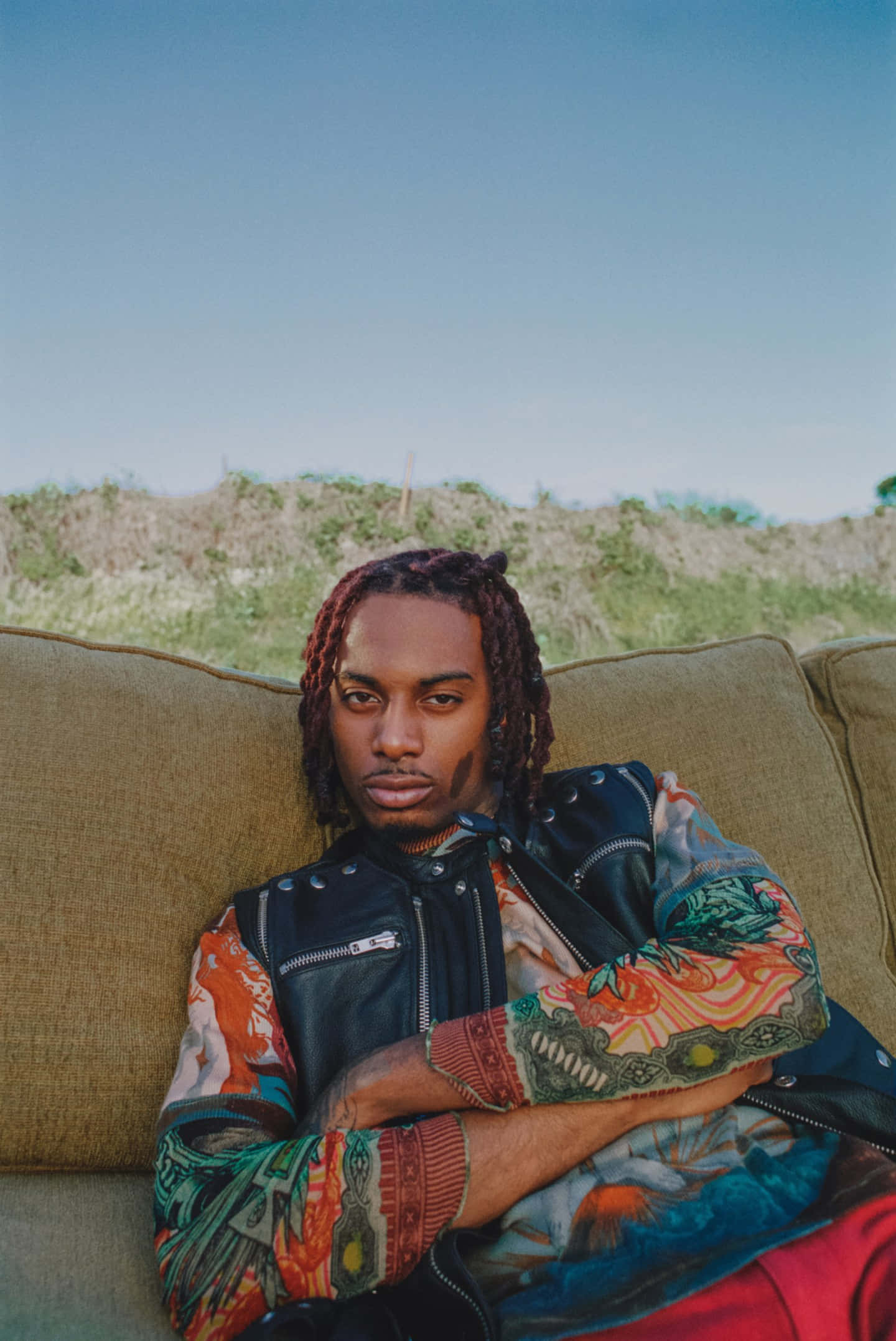 Playboi Carti iPhone 桌布 壁紙 — 一位辮子髮型的年輕男子自信地坐在戶外的棕褐色沙發上，穿著彩色圖案襯衫，外搭黑色皮背心，背景是清澈的藍天。