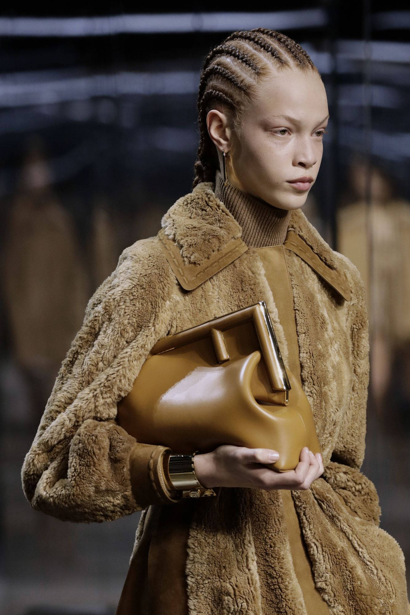 Fendi 桌布 壁紙 — 一位辮子髮型的模特兒穿著毛絨絨的棕色皮草外套，在昏暗的 runway 燈光下拿著相配的皮革手拿包。