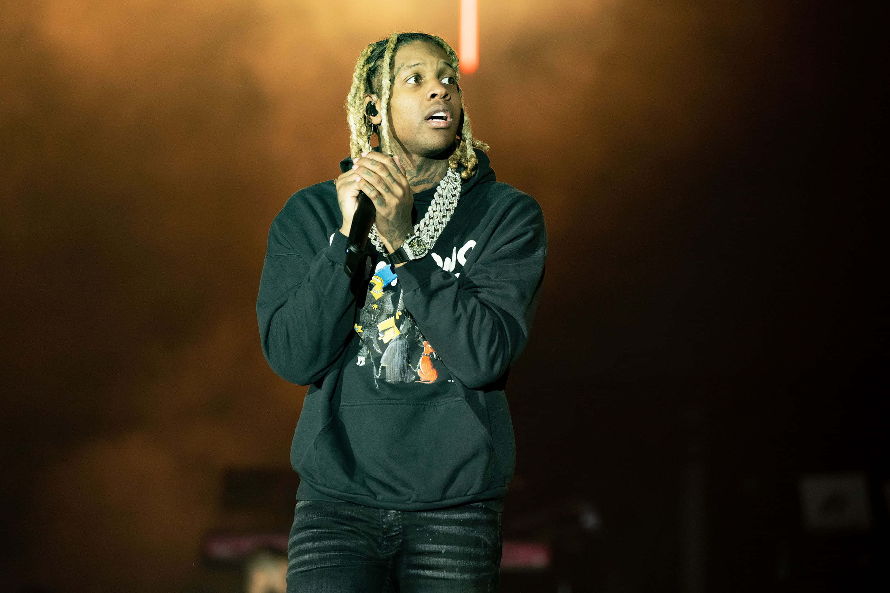 Lil Durk 桌布 壁紙 — 一位辮子髮型的饒舌歌手在溫暖的橙色燈光下拿著麥克風在舞台上表演。