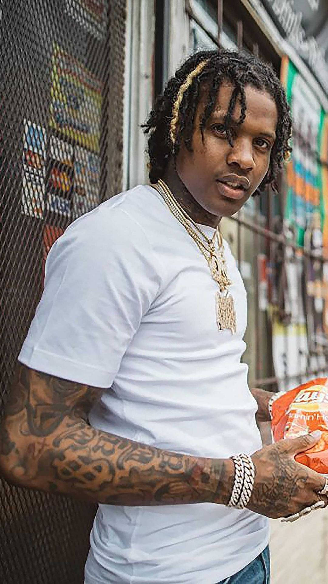 Lil Durk 桌布 壁紙 — 一位辮髮、有刺青的年輕男子在都市小巷中靠著鐵絲網圍欄，手持一包洋芋片。
