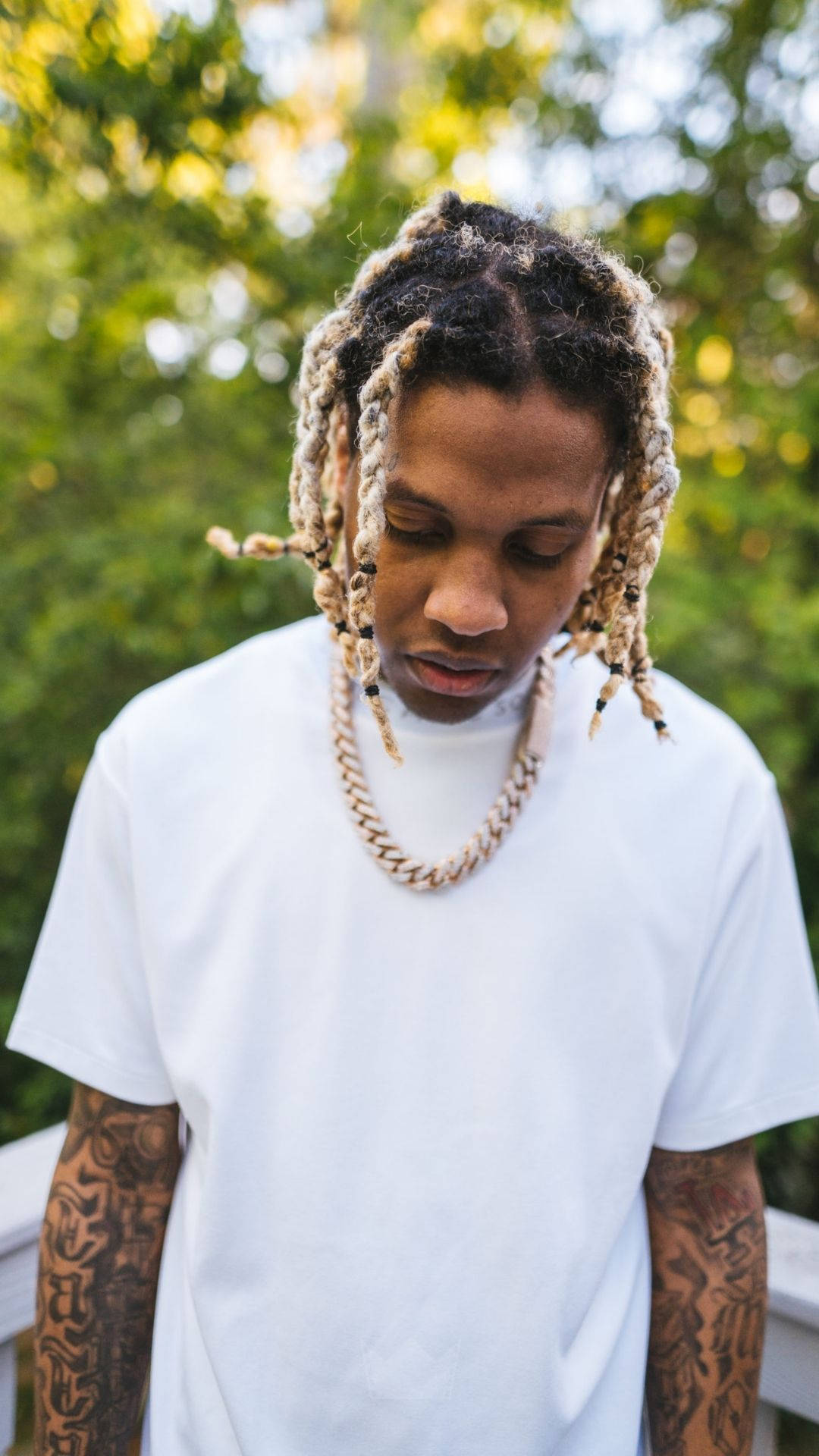 Lil Durk 桌布 壁紙 — 一位辮髮紋身的年輕男子，穿著白色T恤和鏈條項鍊，在陽光普照的綠色背景下低頭凝視。