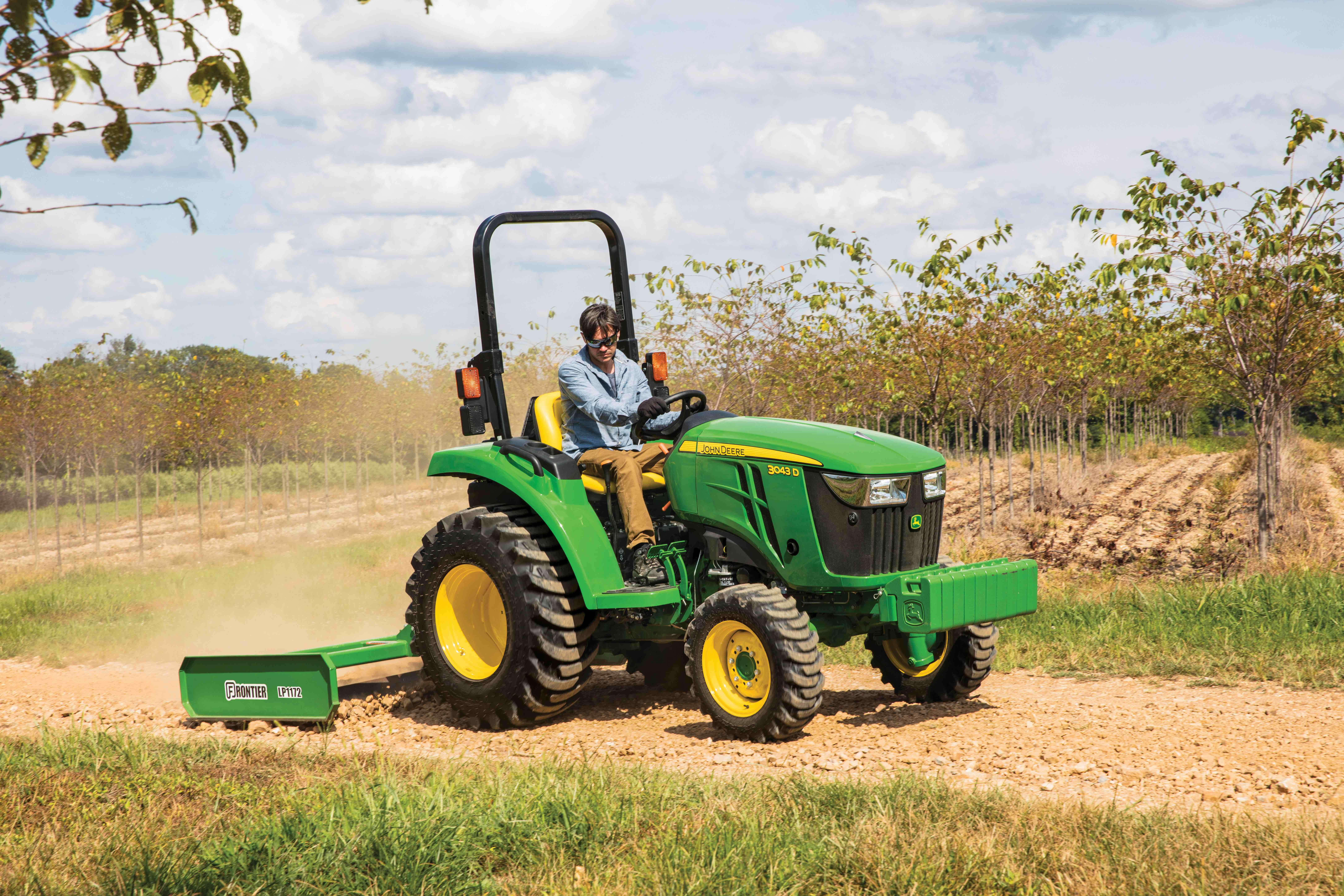 Tractor 桌布 壁紙 — 一位農夫駕駛一輛綠色約翰迪爾拖拉機，黃色車輪，沿著一條塵土飛揚的鄉村小路穿過一片幼樹田，天空部分多雲。