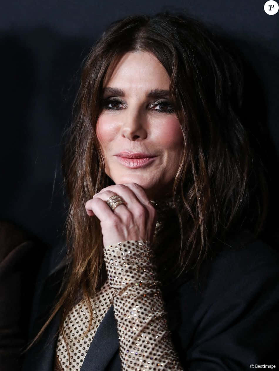 Sandra Bullock 2021 桌布 壁紙 — 一位迷人女性，深色波浪長髮，手托下巴，手上戴有戒指和閃亮袖飾，背景深色。