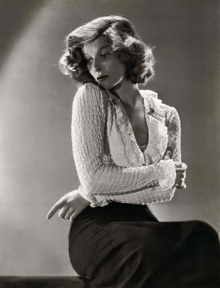 katharine hepburn 桌布 壁紙 — 一位迷人的女士，捲髮，坐在攝影棚中，穿著有質感的襯衫和深色裙子，沐浴在戲劇性的聚光燈下。