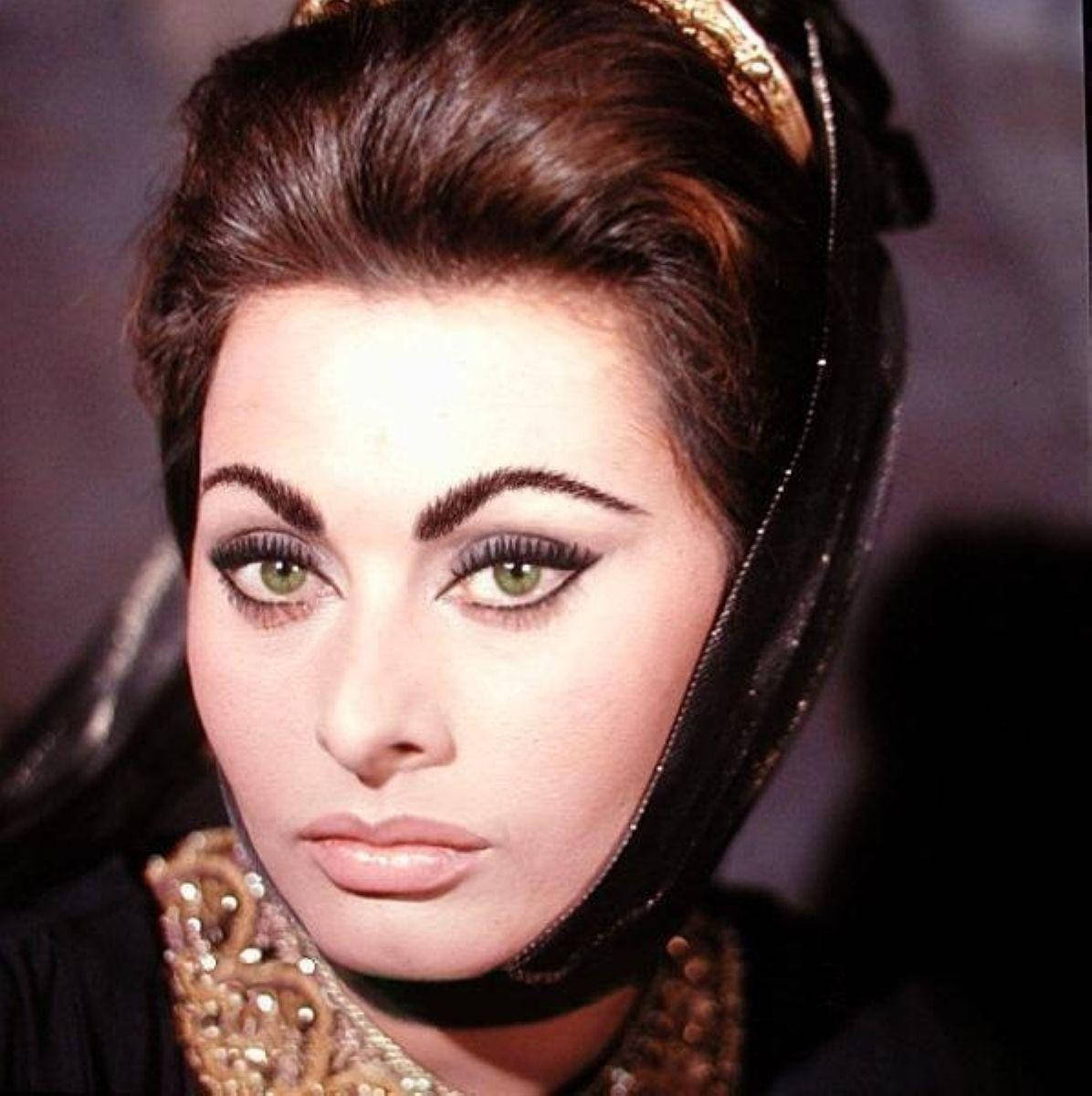 Sophia Loren 桌布 壁紙 — 一位迷人的女性，擁有引人注目的綠色眼睛和大膽的黑色眼線，戴著華麗的金色頭飾和珠寶項圈，凝視著。