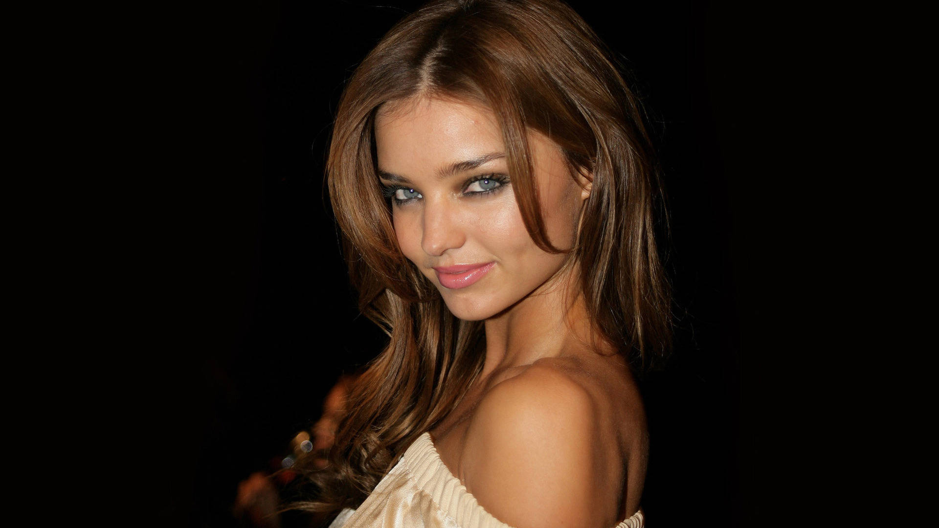 Miranda Kerr 桌布 壁紙 — 一位迷人的女性，長長的棕色頭髮和引人注目的藍色眼睛，在黑色背景前溫柔地微笑。