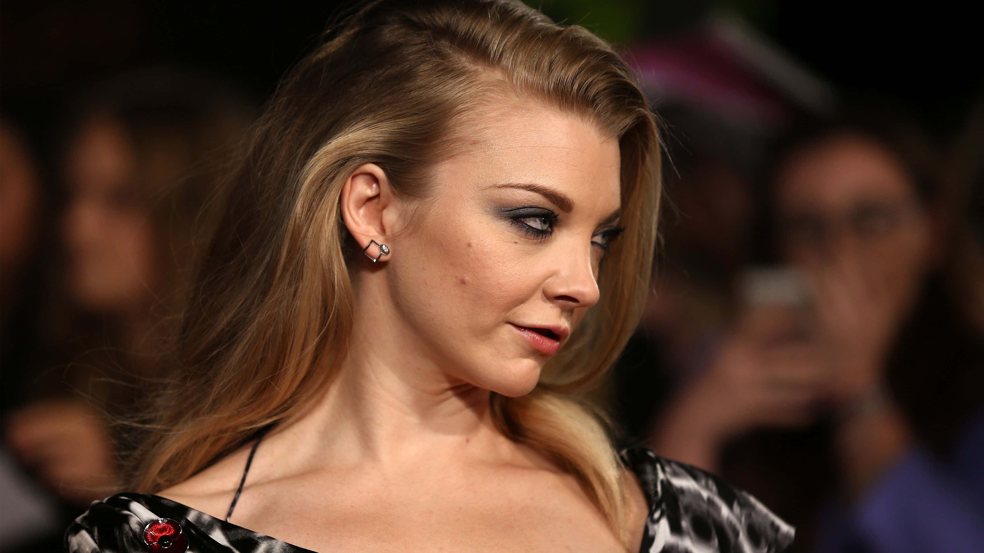 natalie dormer 桌布 壁紙 — 一位迷人的金髮女郎，化著煙燻眼妝，穿著黑色圖案連衣裙，在擁擠的活動中側視。
