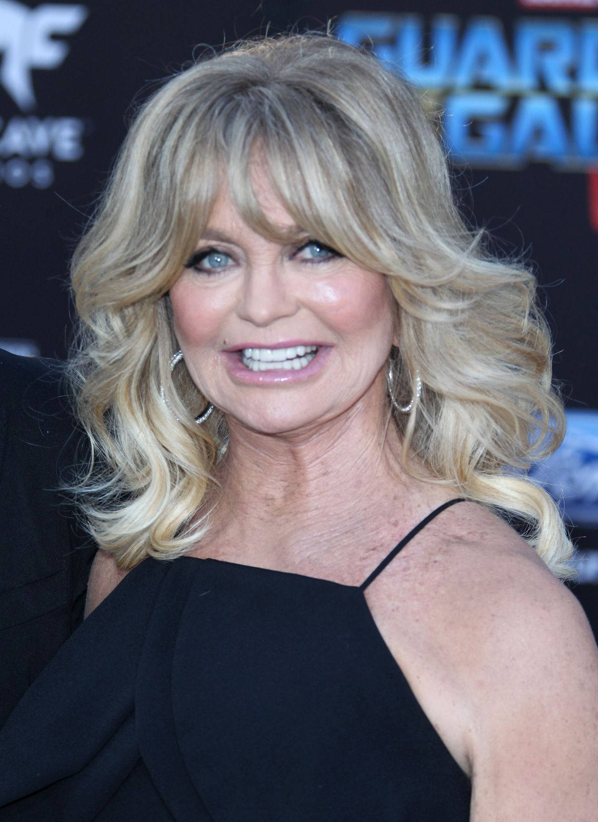 Celebrity 桌布 壁紙 — 一位迷人的金髮女郎，波浪捲髮和明亮的藍眼睛，穿著黑色連衣裙和銀色耳環對著鏡頭微笑。