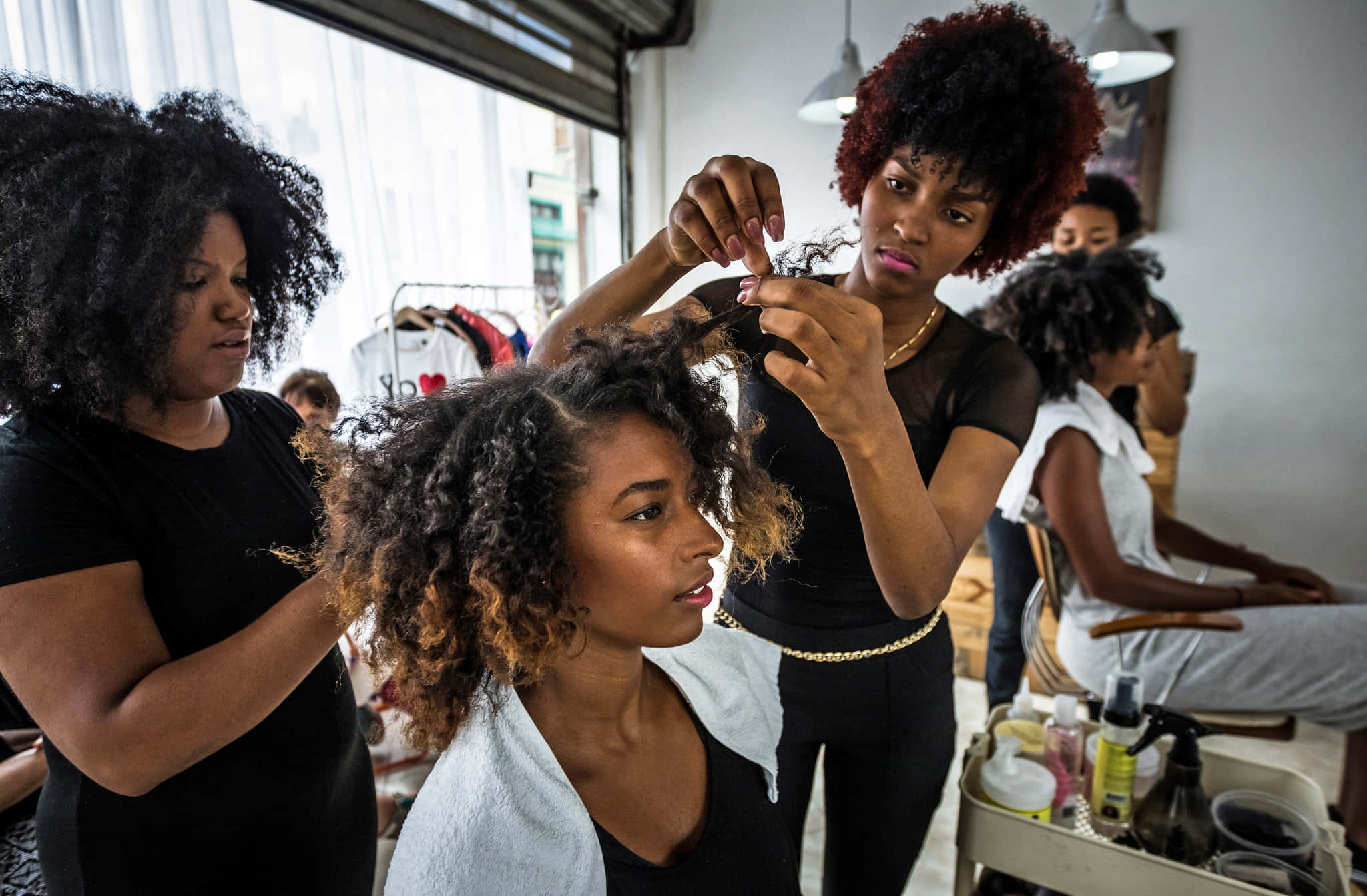 Hair Salon 桌布 壁紙 — 一位造型師在光線充足的沙龍裡，自然光透過大窗戶灑進來，小心翼翼地為客戶編織捲髮。